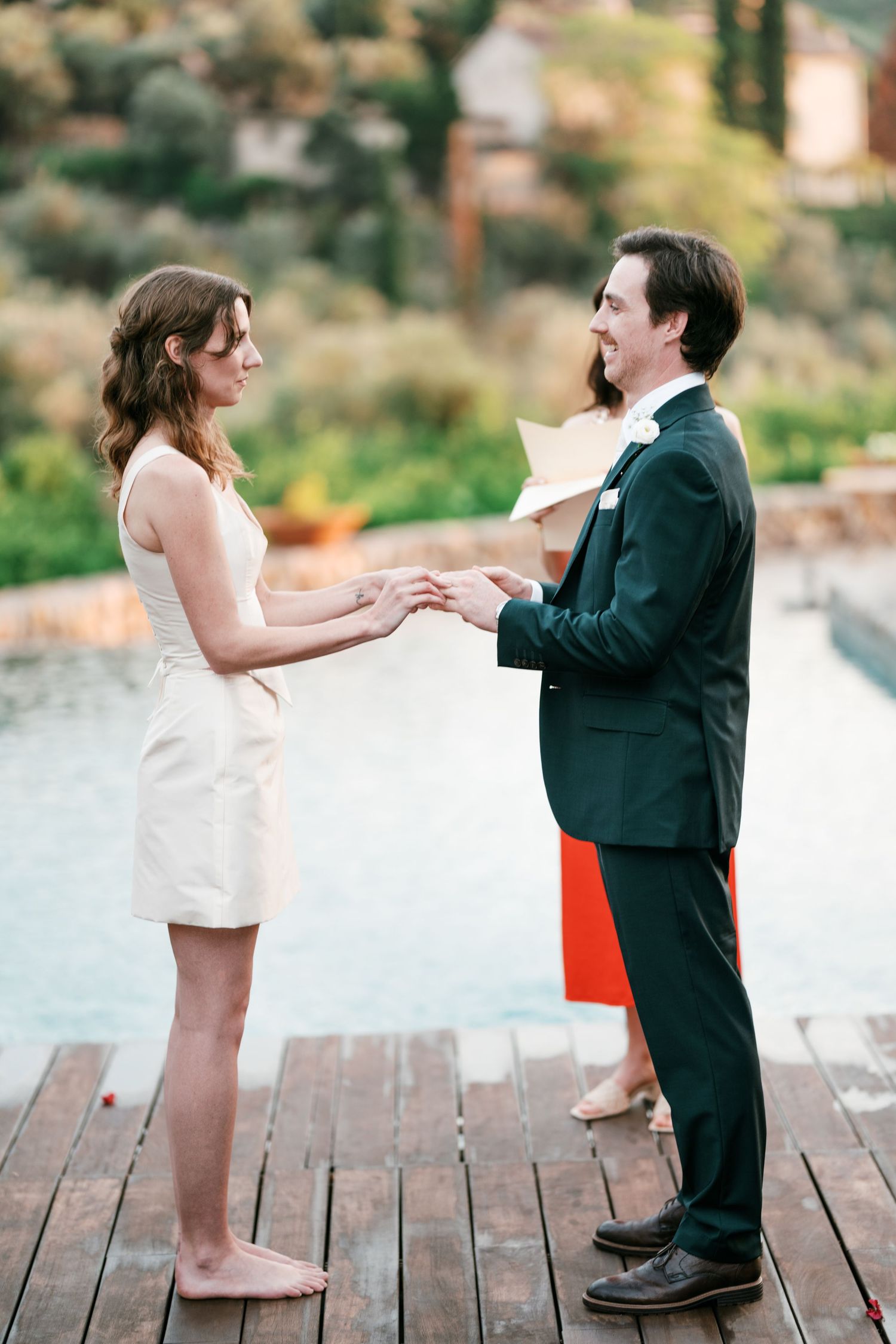 Bride in a short white dress and groom in a dark suit hold hands during an outdoor wedding ceremony on a wooden deck by a pool.