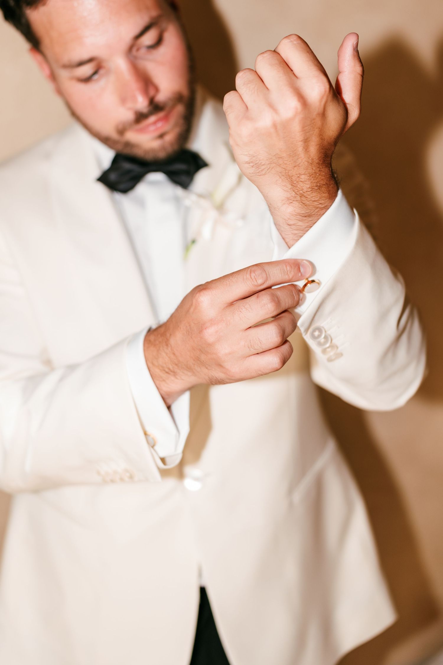 A groom in a white tuxedo jacket and black bow tie fastens his cufflinks while preparing for his wedding.