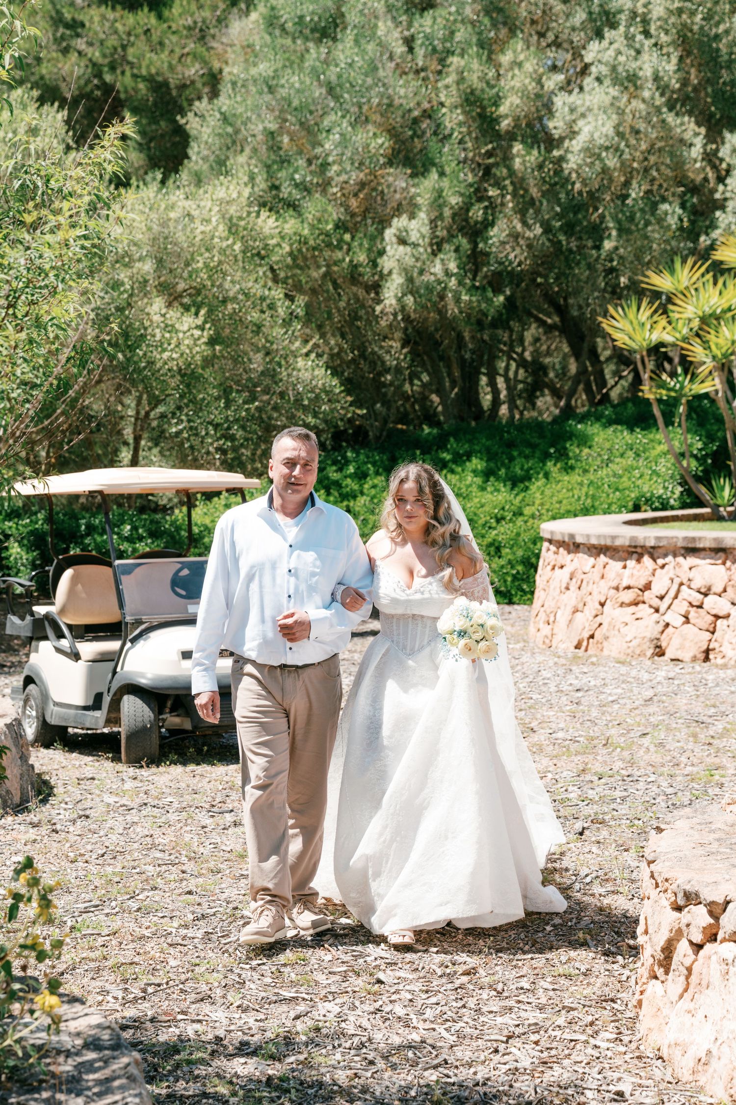 Bride in white gown walks with man in white shirt through lush garden, golf cart visible in background in Mallorca.