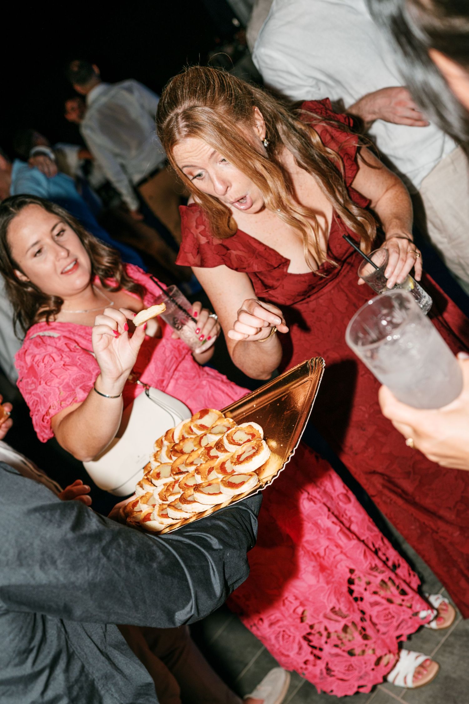 Two women in red and pink dresses laugh at a festive party while holding drinks and sampling appetizers from a golden serving tray.