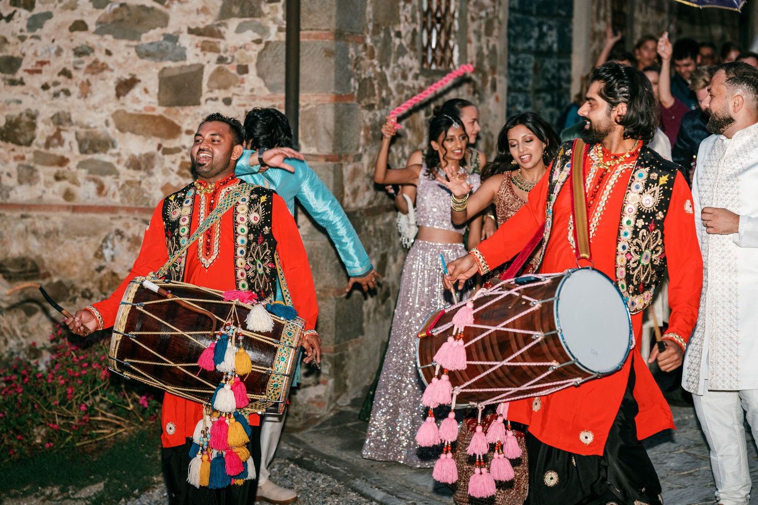 Two dhol drummers in red outfits with colorful tassels lead a lively wedding procession as guests dance outside an old stone building.