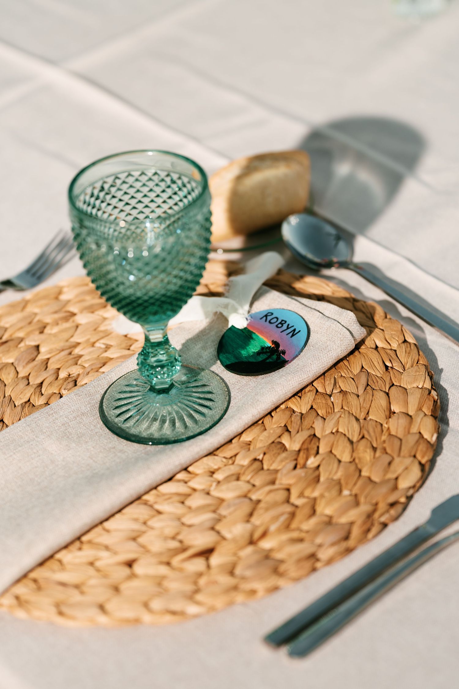 Green textured wine glass on a woven rattan placemat with a linen napkin, name tag, bread, and silverware on a white tablecloth.