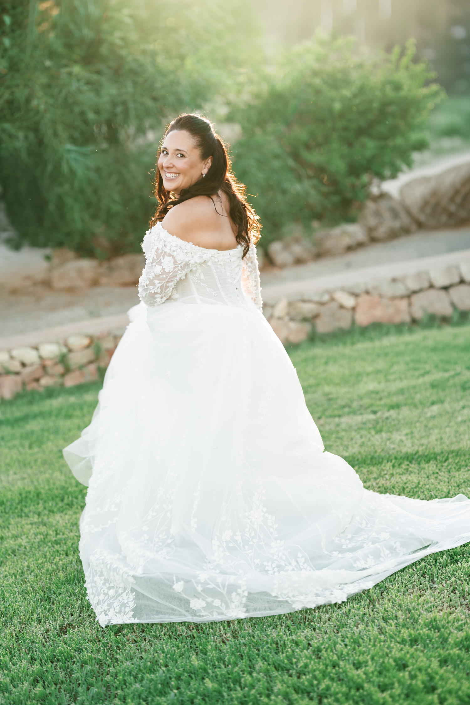 Smiling bride in an off-the-shoulder white lace wedding gown looks back over her shoulder while standing on a sunlit grassy lawn.