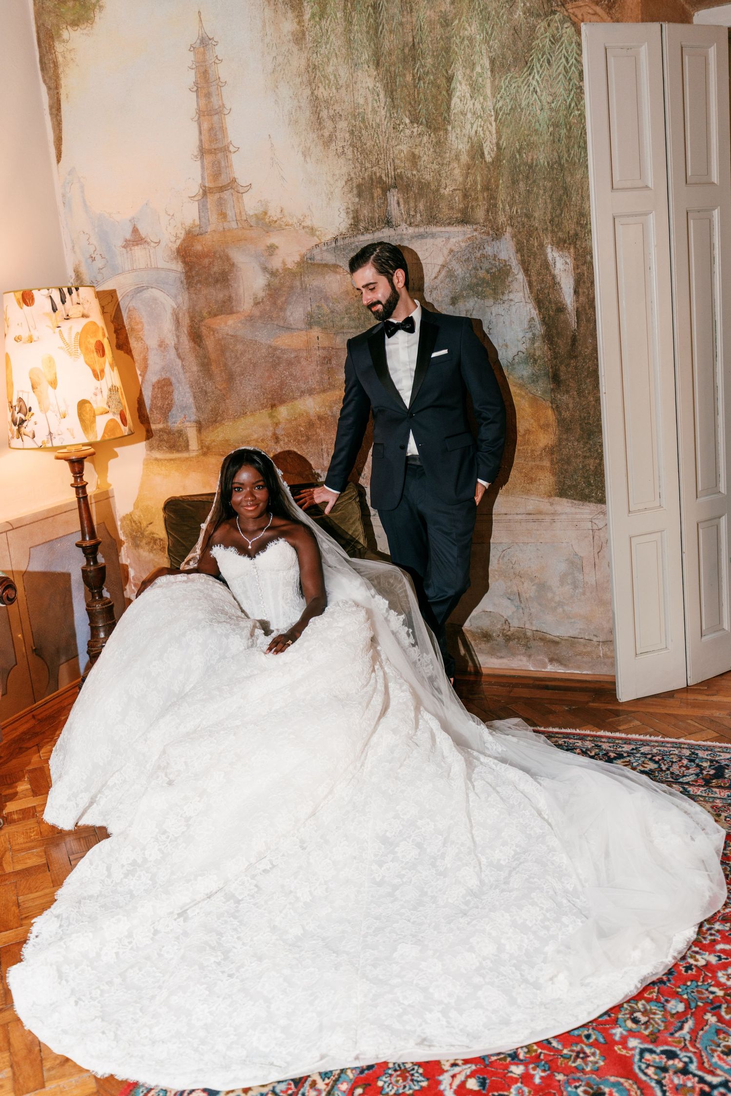 Bride in a voluminous white ball gown with a long veil sits in an armchair beside a groom in a black tuxedo, in front of an antique mural.