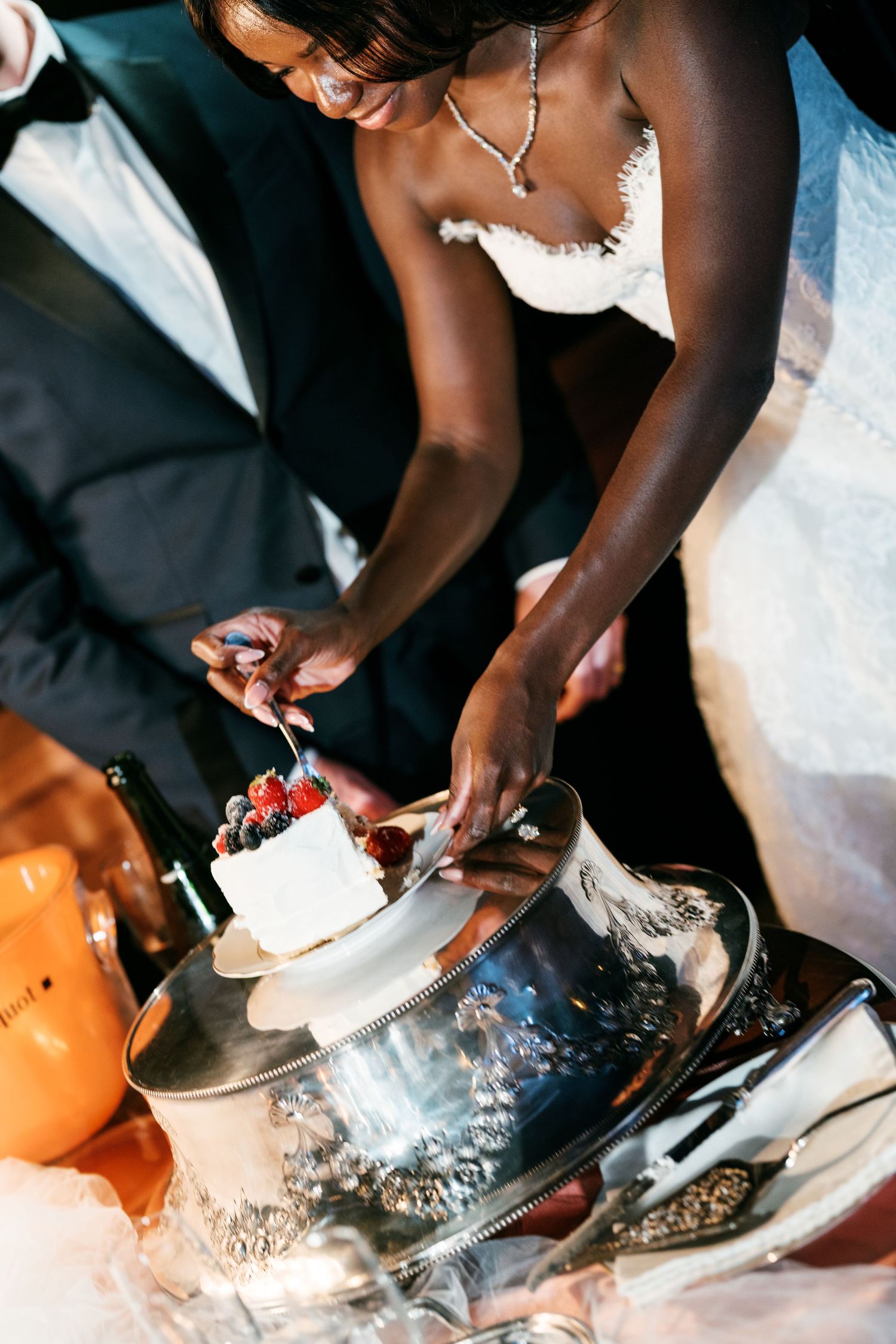 Bride in a lace wedding dress and pearl necklace cuts a berry-topped wedding cake on a silver stand beside the groom.