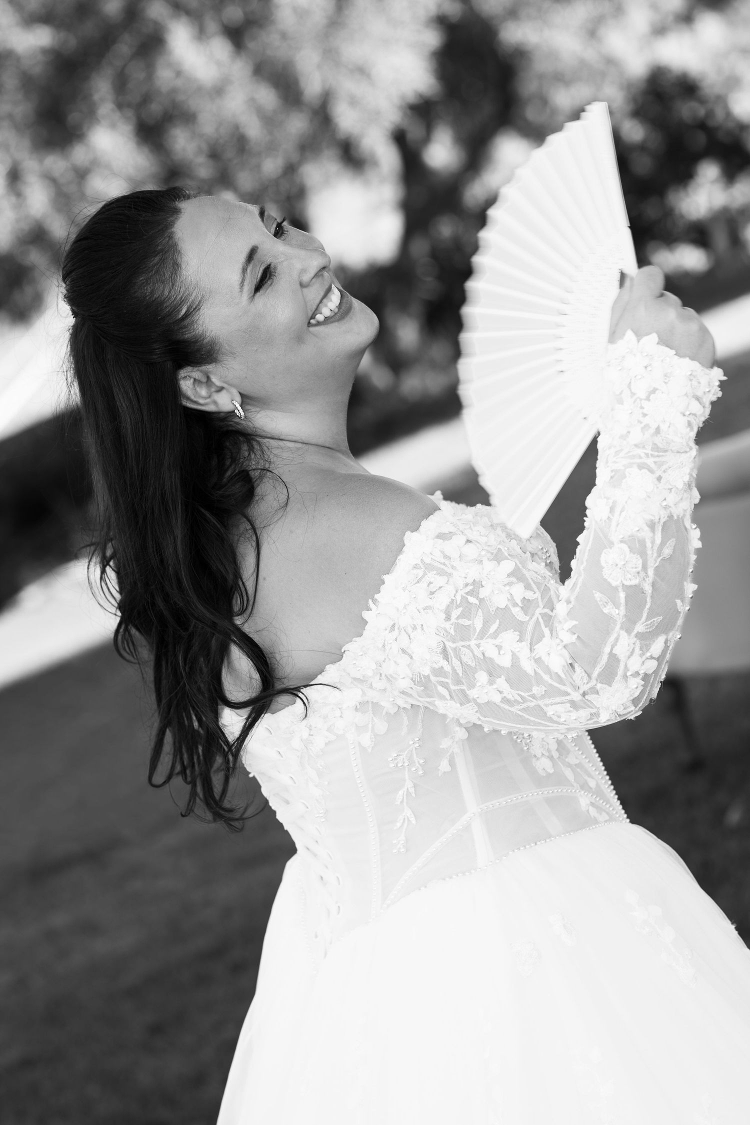 Black and white photo of a laughing bride in a lace off-the-shoulder wedding gown holding a folding fan outdoors.