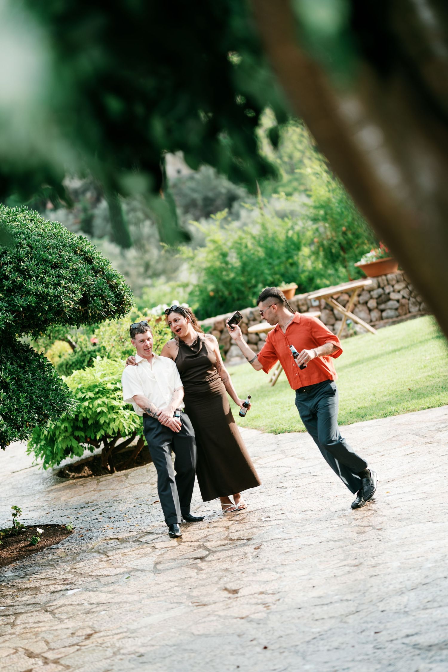 Three guests walk and laugh along a stone path in a sunny garden with lush greenery at an outdoor event.