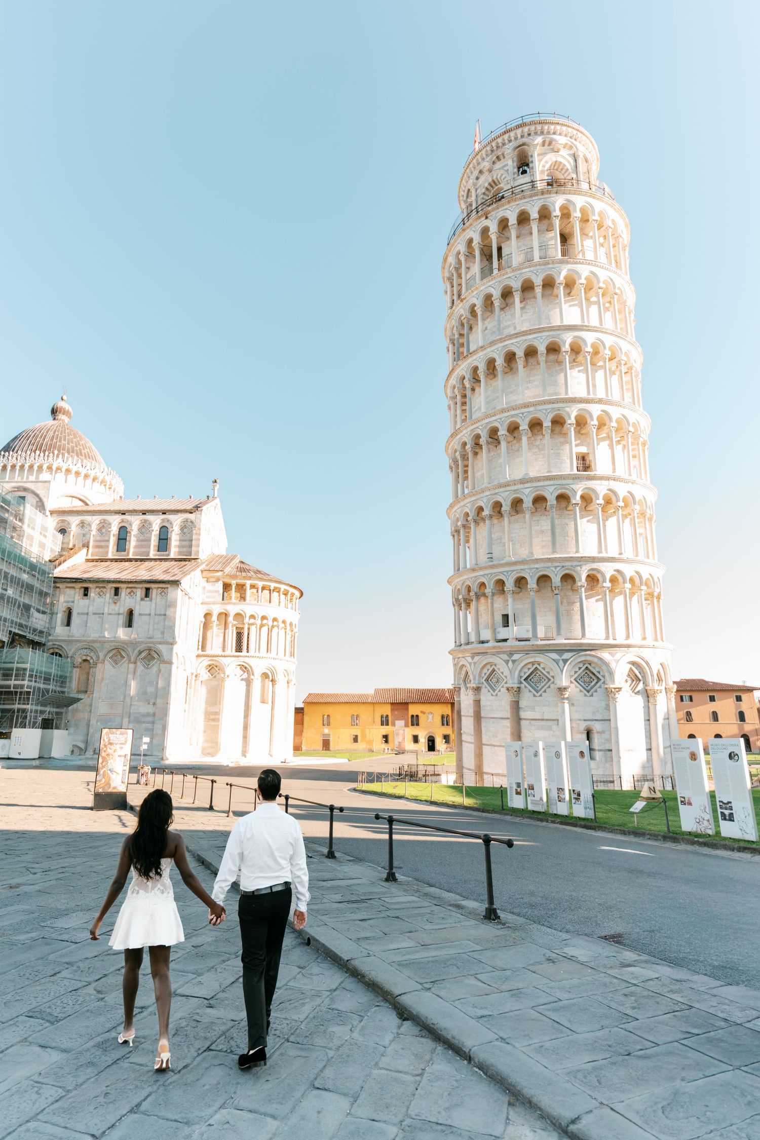 Couple in white dress and shirt walk hand in hand toward the Leaning Tower of Pisa and cathedral on a sunny day in Italy.