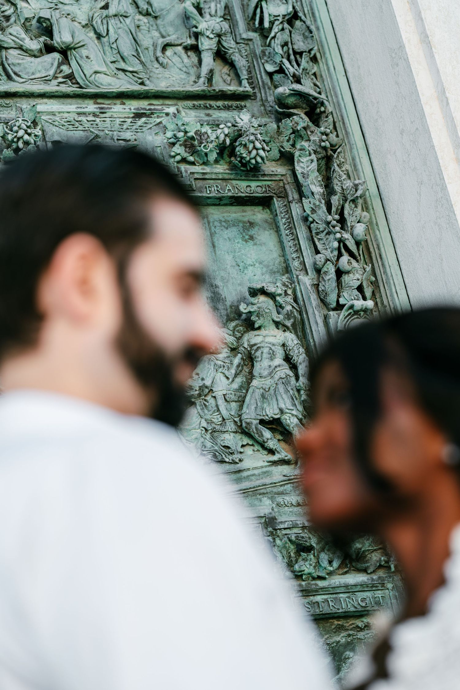 Blurred couple smiling at each other in front of an ornate weathered bronze church door with detailed relief sculptures.