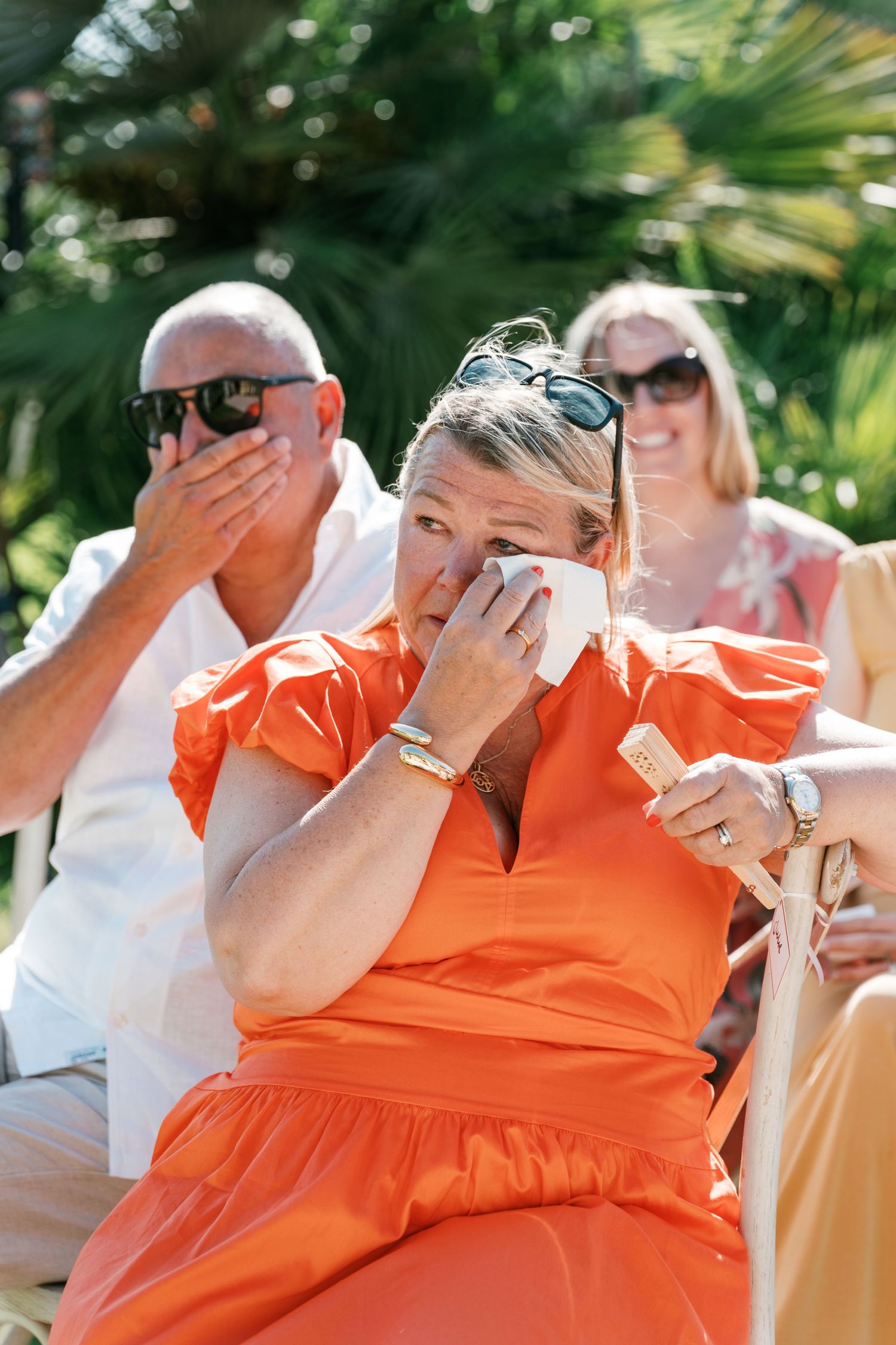 Emotional wedding guest in orange dress wiping tears with tissue during outdoor ceremony on sunny day. Mallorca wedding photographer