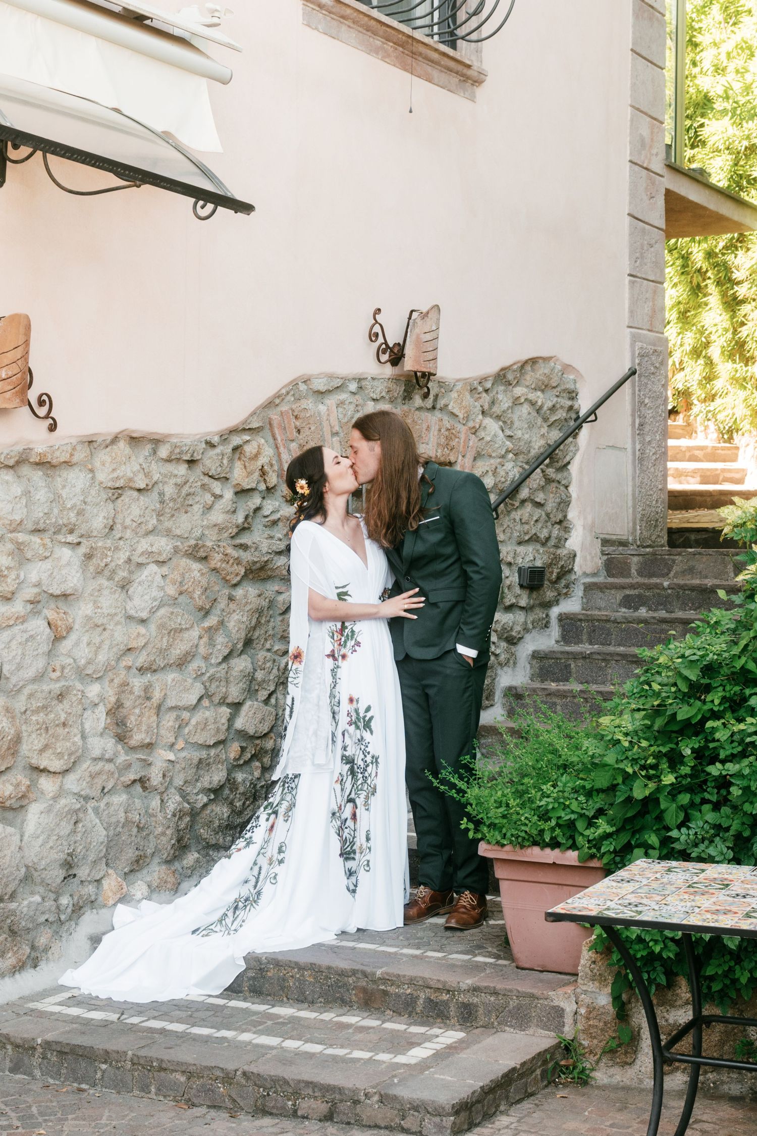 Bride and groom share a romantic kiss on stone steps beside a rustic villa wall with greenery. Tuscany wedding photographer, filmmaker