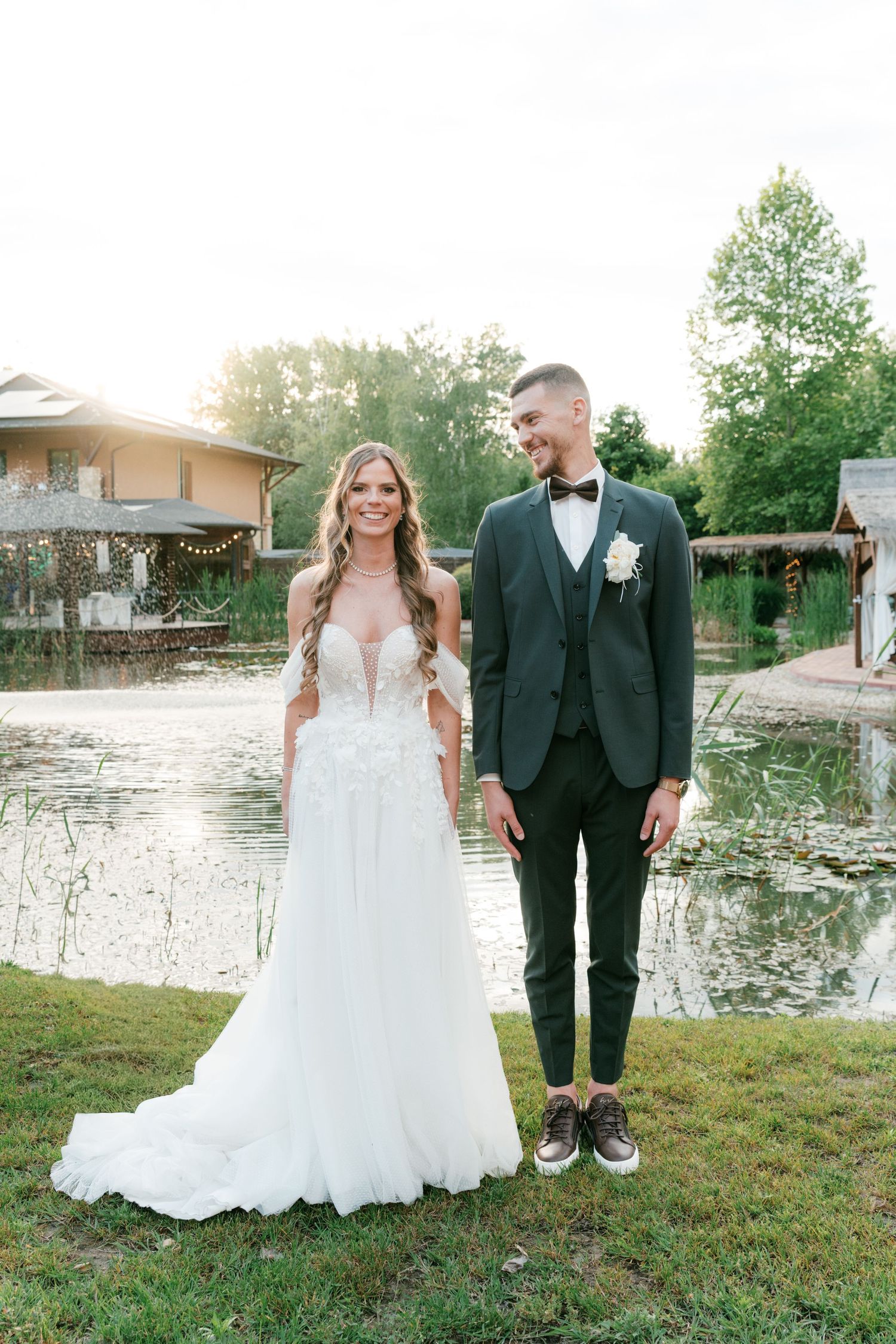 Smiling bride and groom stand together by a reflective pond at sunset wedding venue. Mallorca wedding photographer and filmmaker.