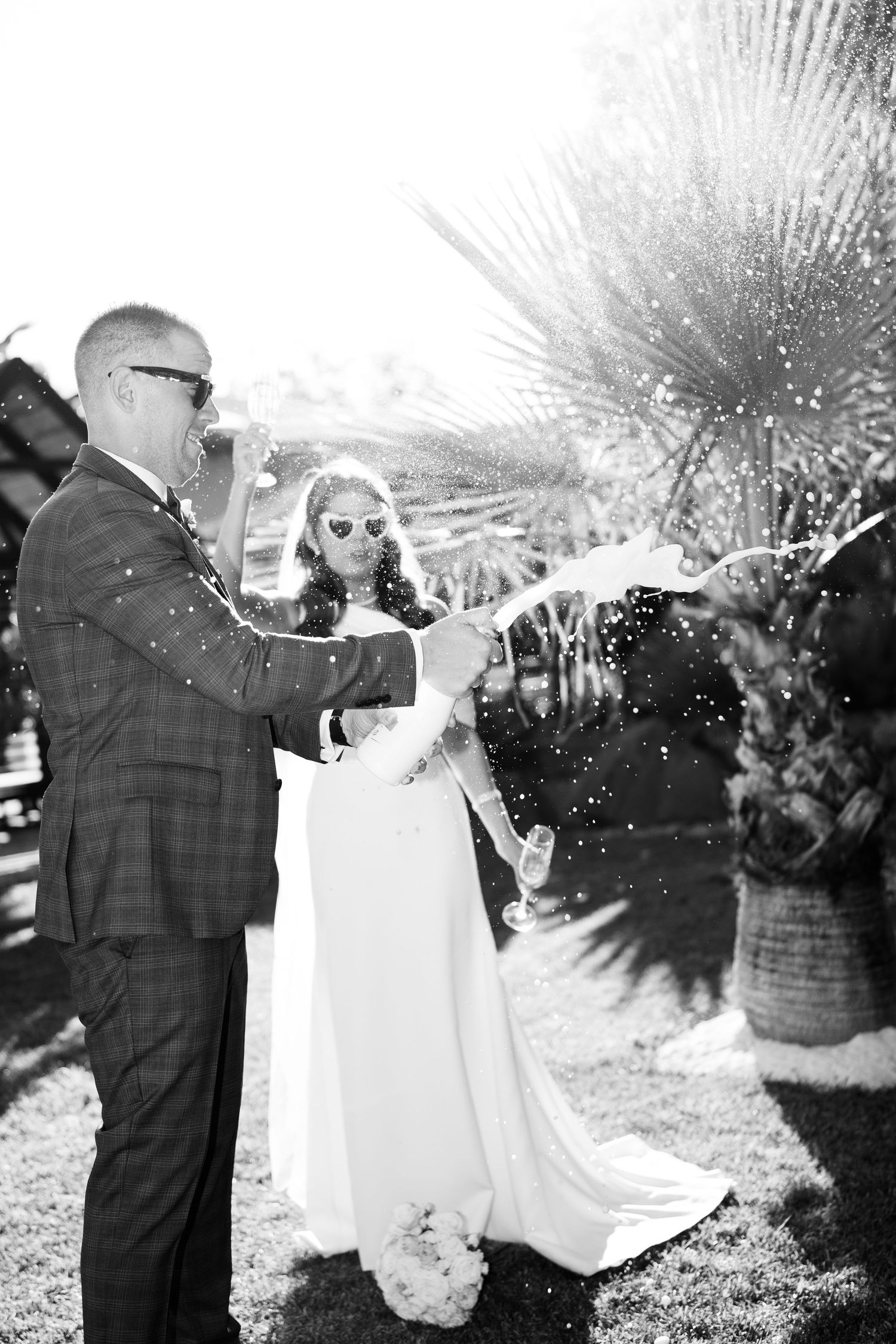 Black and white photo of a bride and groom sabering champagne outdoors, wearing sunglasses, with palm trees in background.
