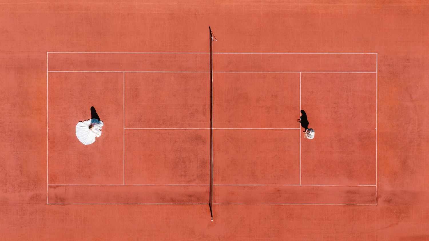 Aerial view of two tennis players on a red clay court, casting long shadows on the orange surface.