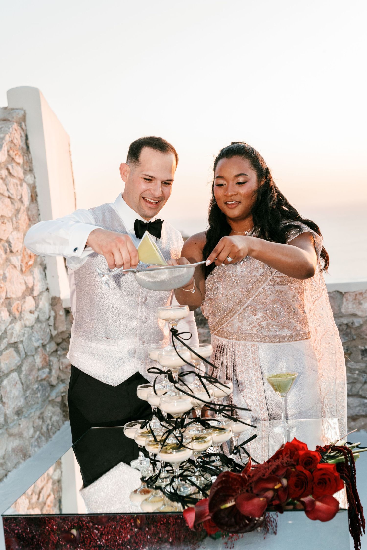 Bride and groom cutting a tiered wedding cake outdoors at sunset, dressed in elegant formal attire.
