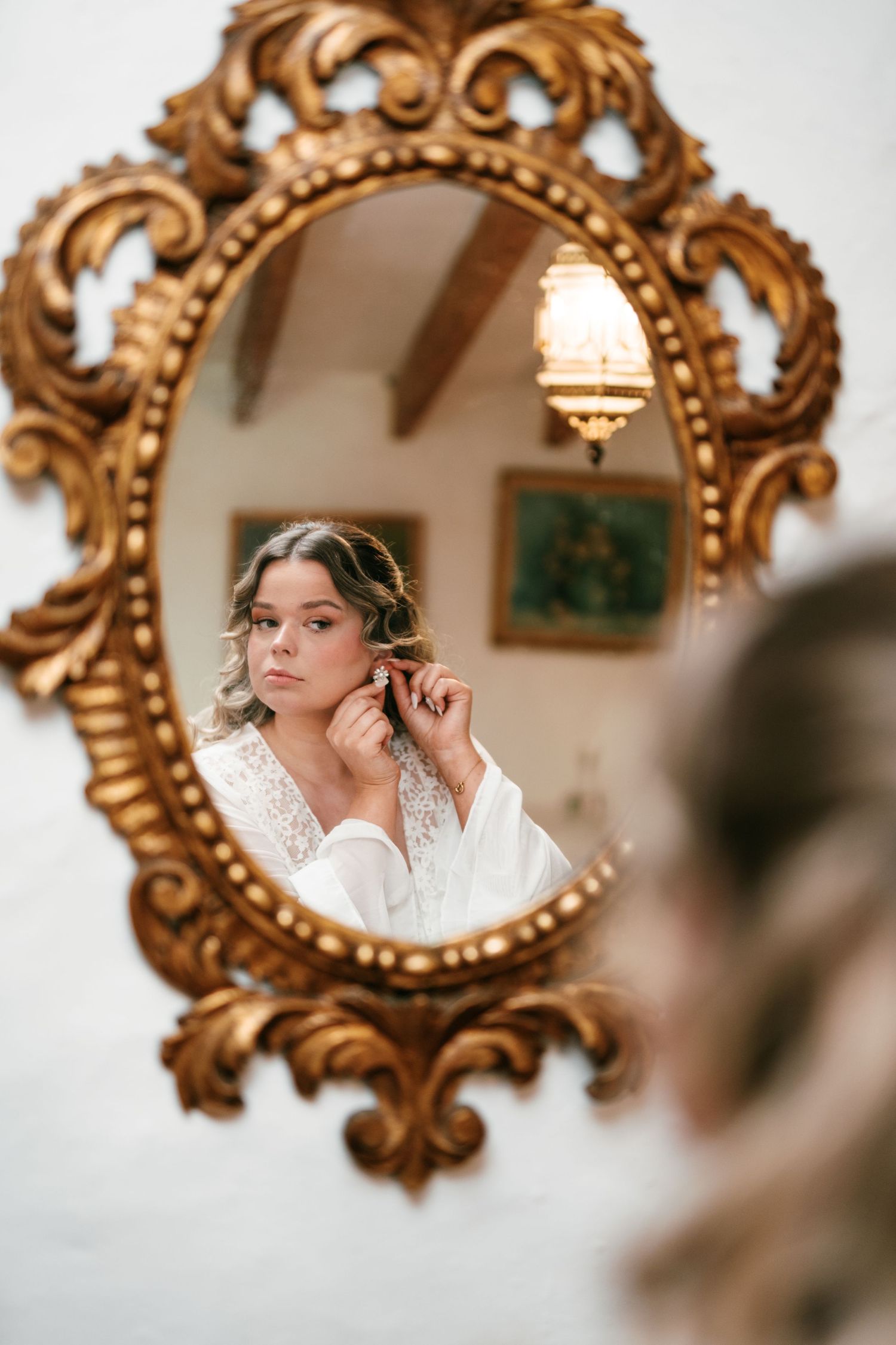 Bride in white robe putting on earrings reflected in ornate gold baroque mirror in elegant room in Mallorca.
