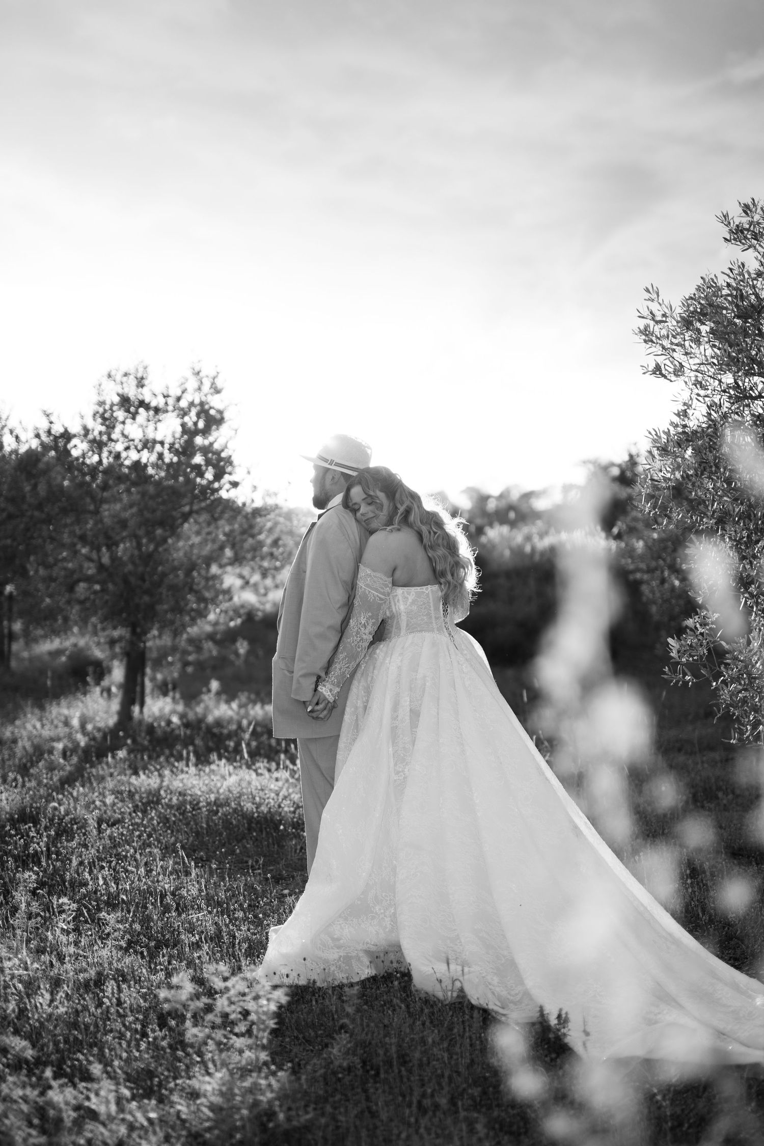 Black and white photo of bride and groom holding hands walking through olive grove at sunset.