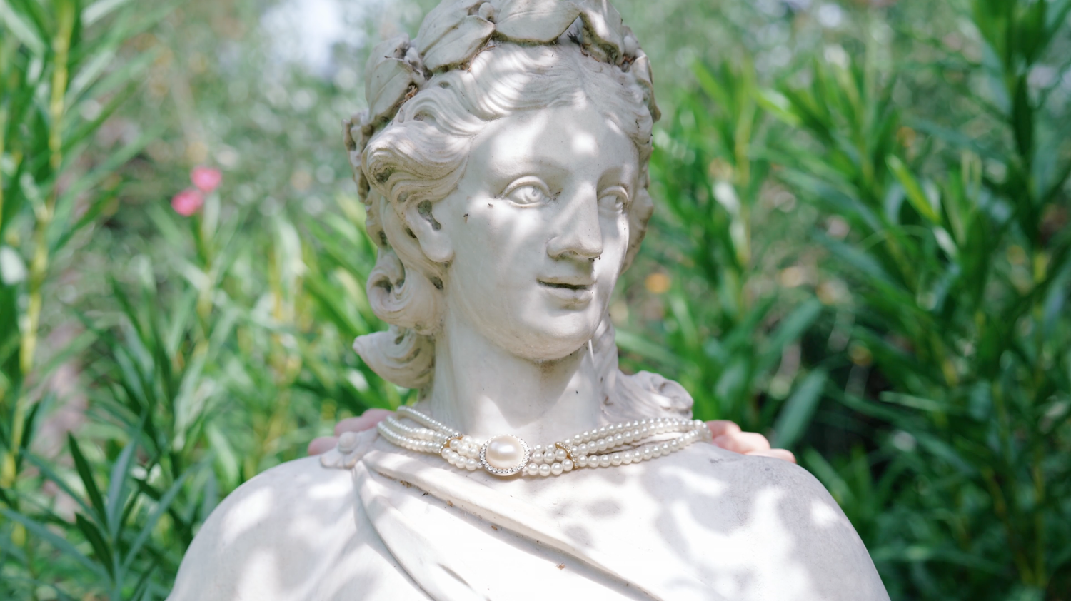 White marble garden statue of a woman with a laurel wreath and pearl necklace, surrounded by green oleander leaves and pink blossoms.