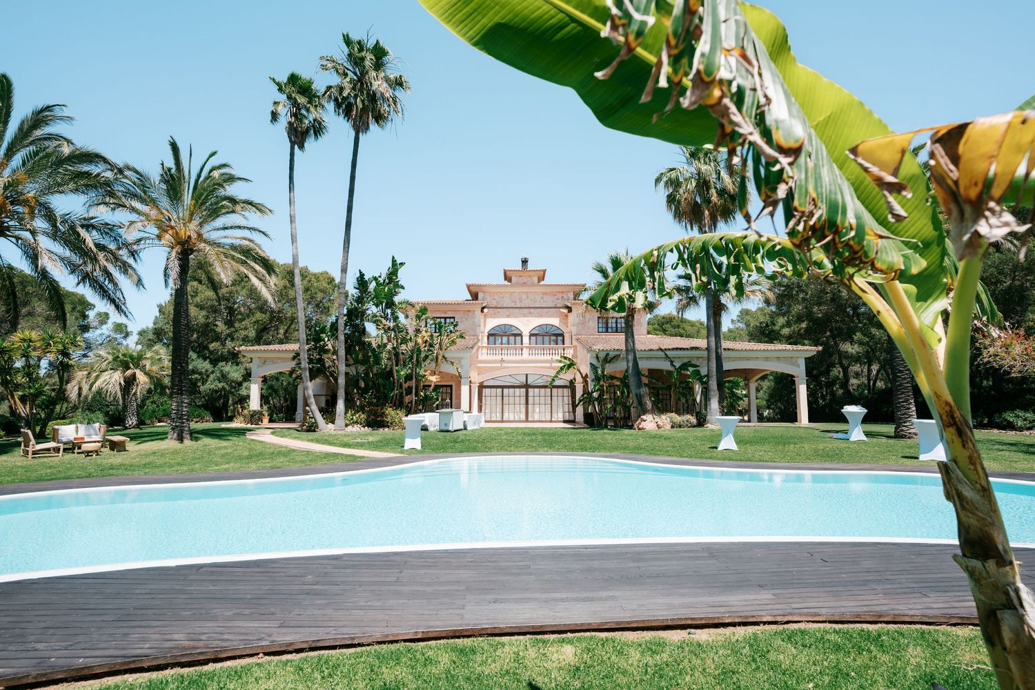 Luxury villa with large pool surrounded by palm trees and lush green gardens on a sunny day.