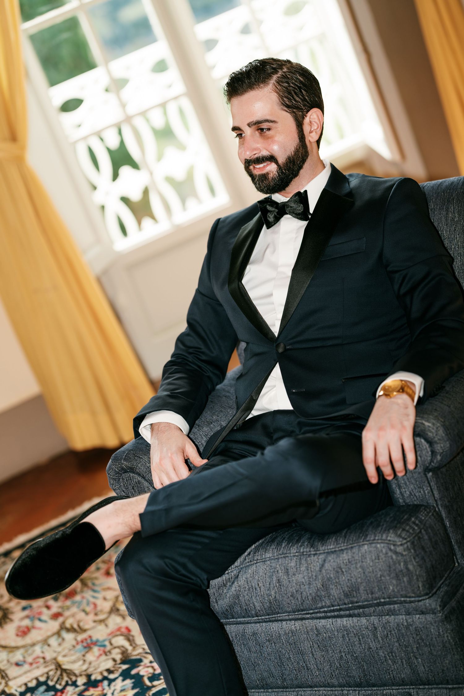 Smiling bearded groom in a black tuxedo and bow tie sits in a gray armchair beside sunlit windows with golden curtains.