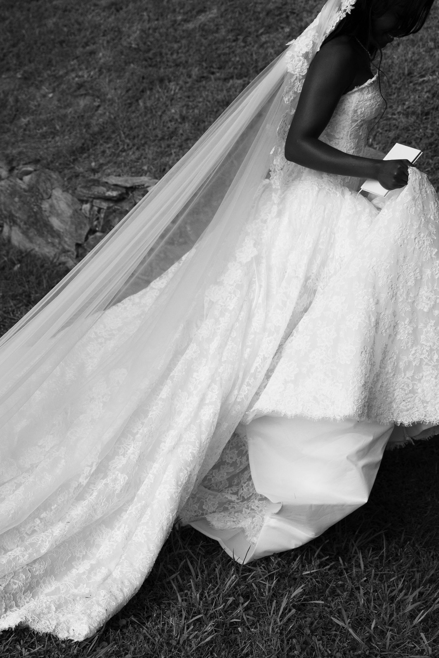 Black and white photo of a bride in a lace wedding gown with a long sheer veil walking across grass while holding a small book.