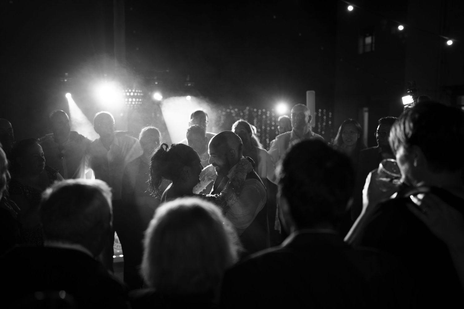 Black and white photo of a lively wedding reception with guests dancing and celebrating under bright stage lights at night.