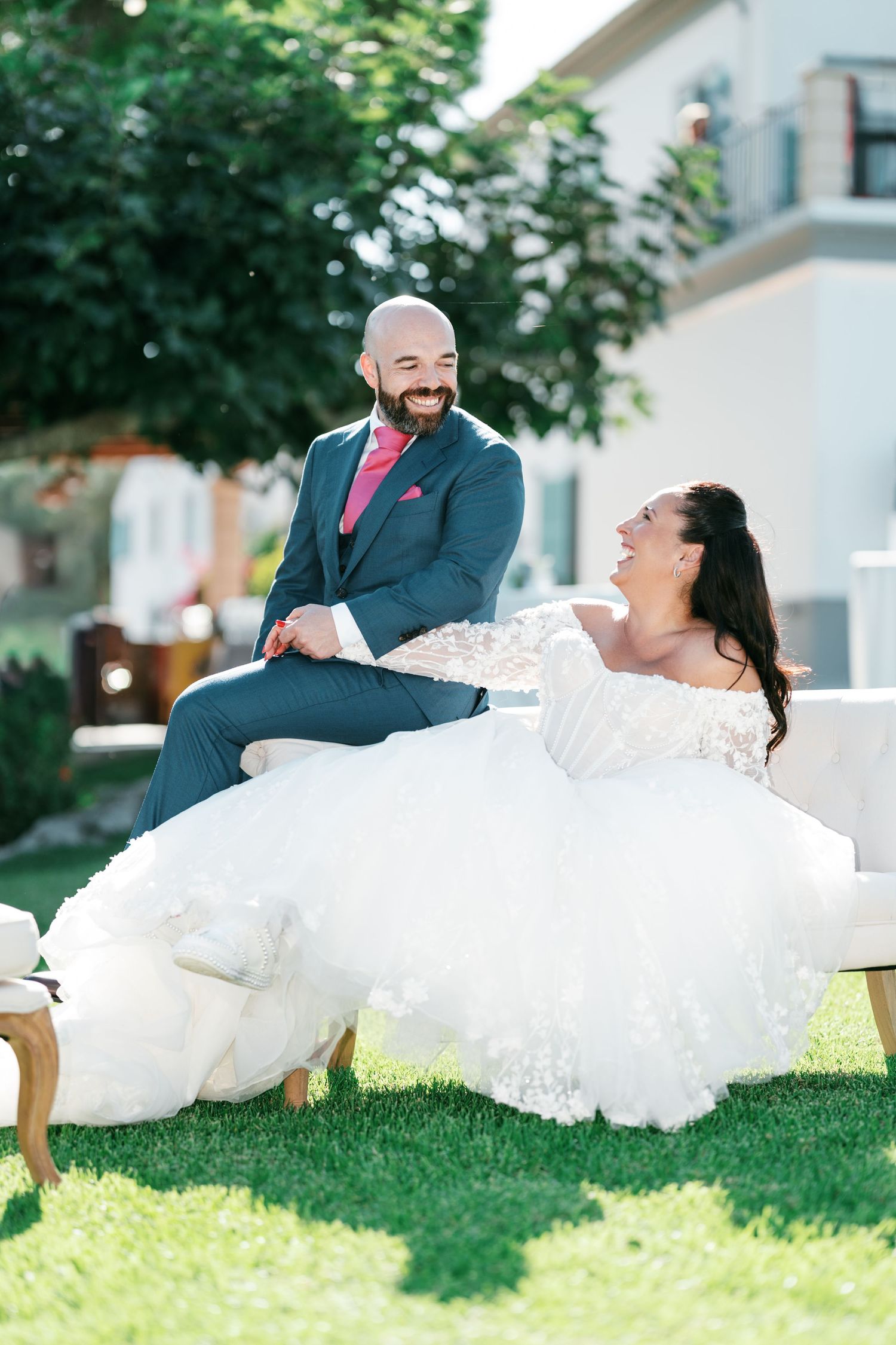 Smiling bride in a lace off-the-shoulder wedding gown laughs with her bearded groom in a blue suit on a sunny green lawn.