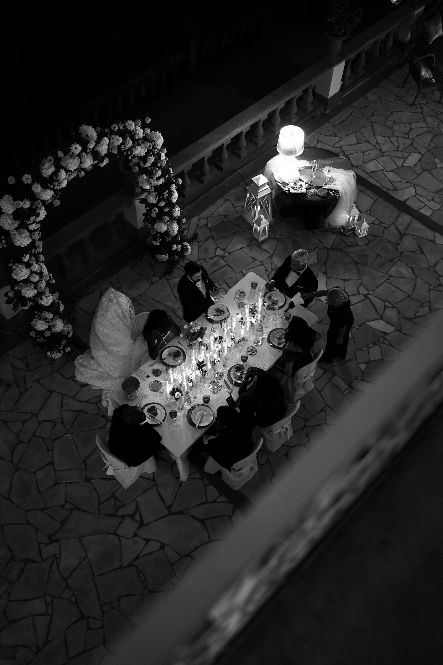 Black and white overhead view of a candlelit wedding dinner table with guests and a bride beside a floral arch on a stone terrace.