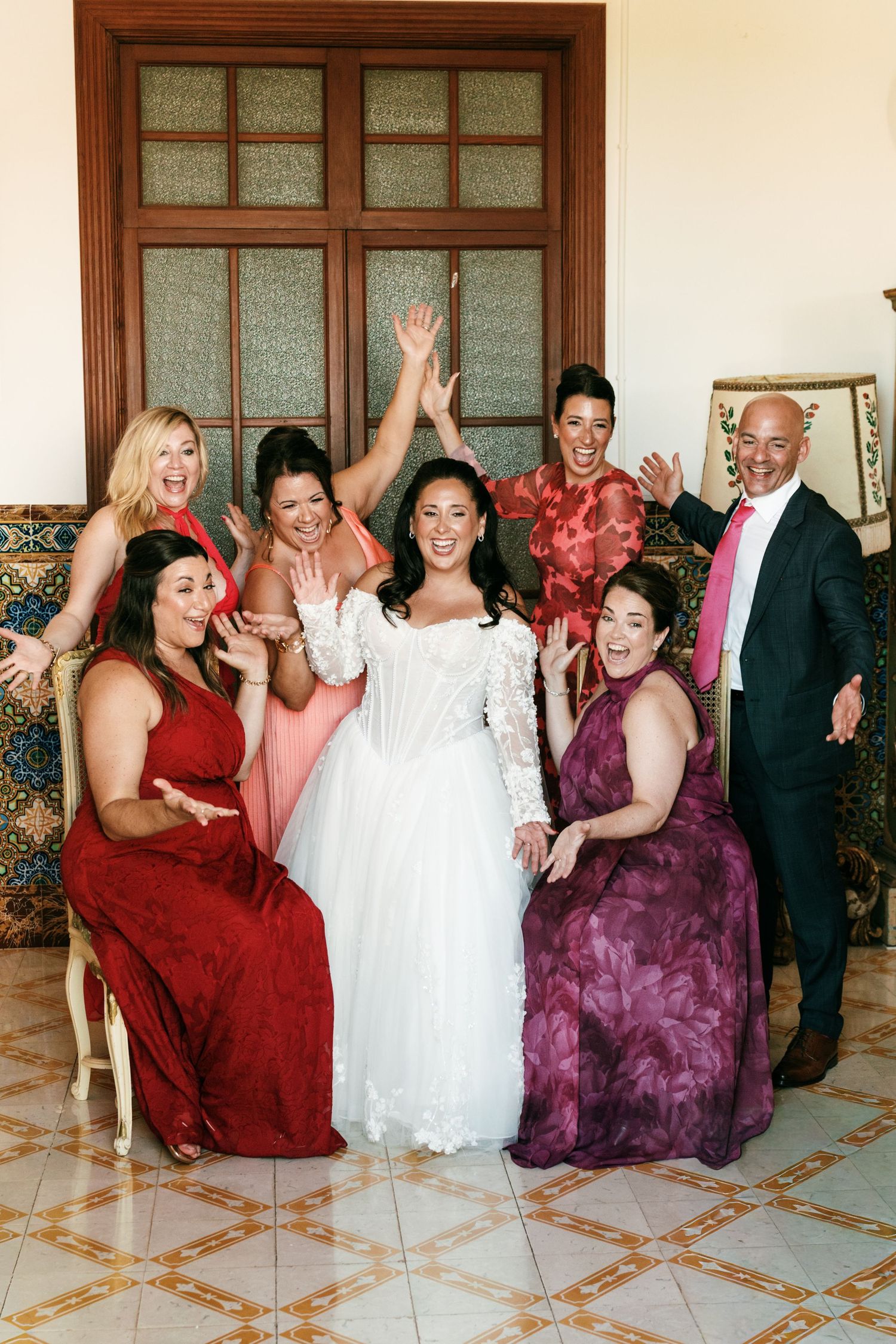 Bride in a white tulle gown poses joyfully with four bridesmaids in red and burgundy dresses and a groomsman in a navy suit.
