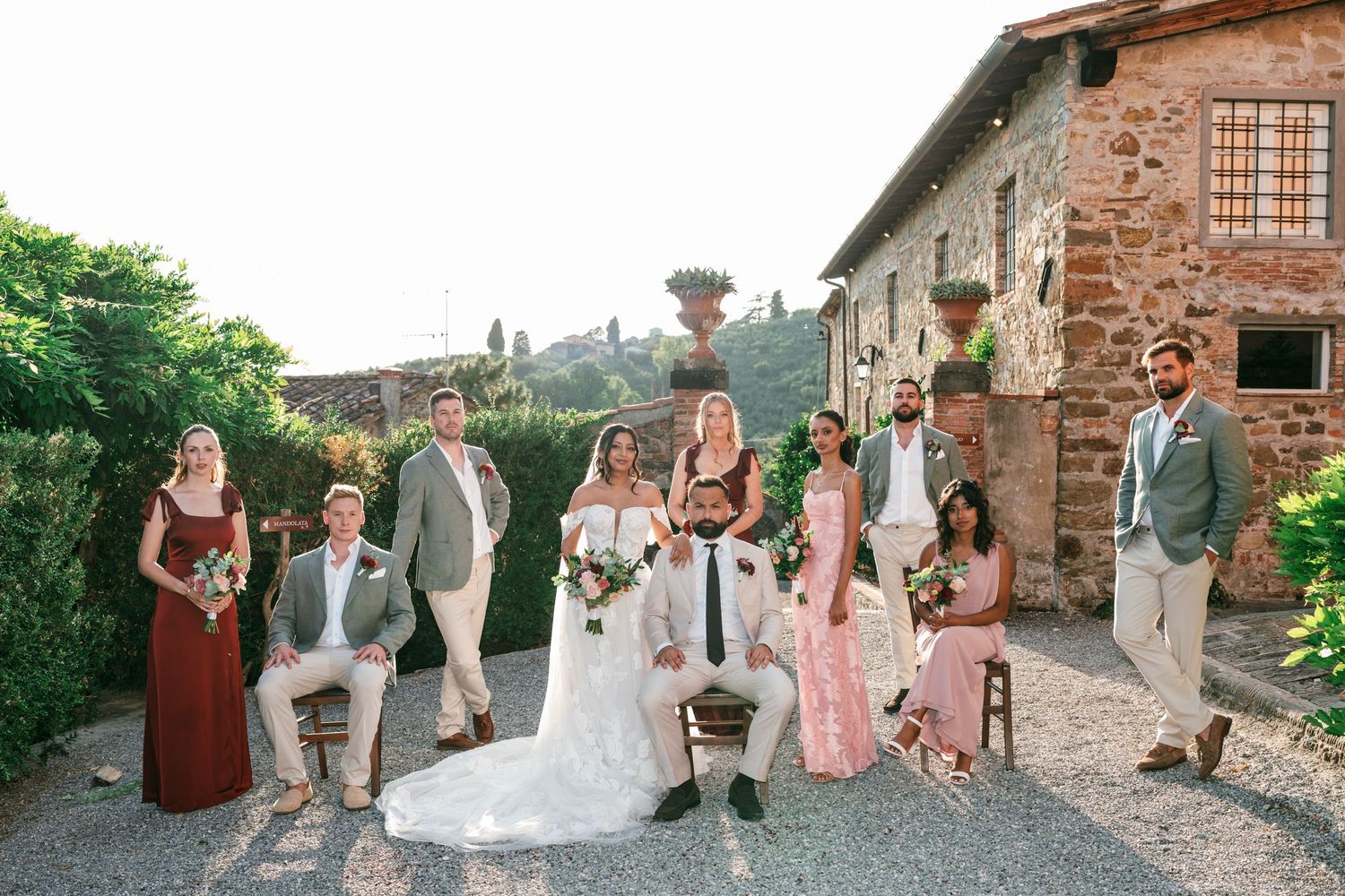 Bride and groom pose with their wedding party in rust and blush dresses and tailored suits outside a rustic Tuscan stone villa.