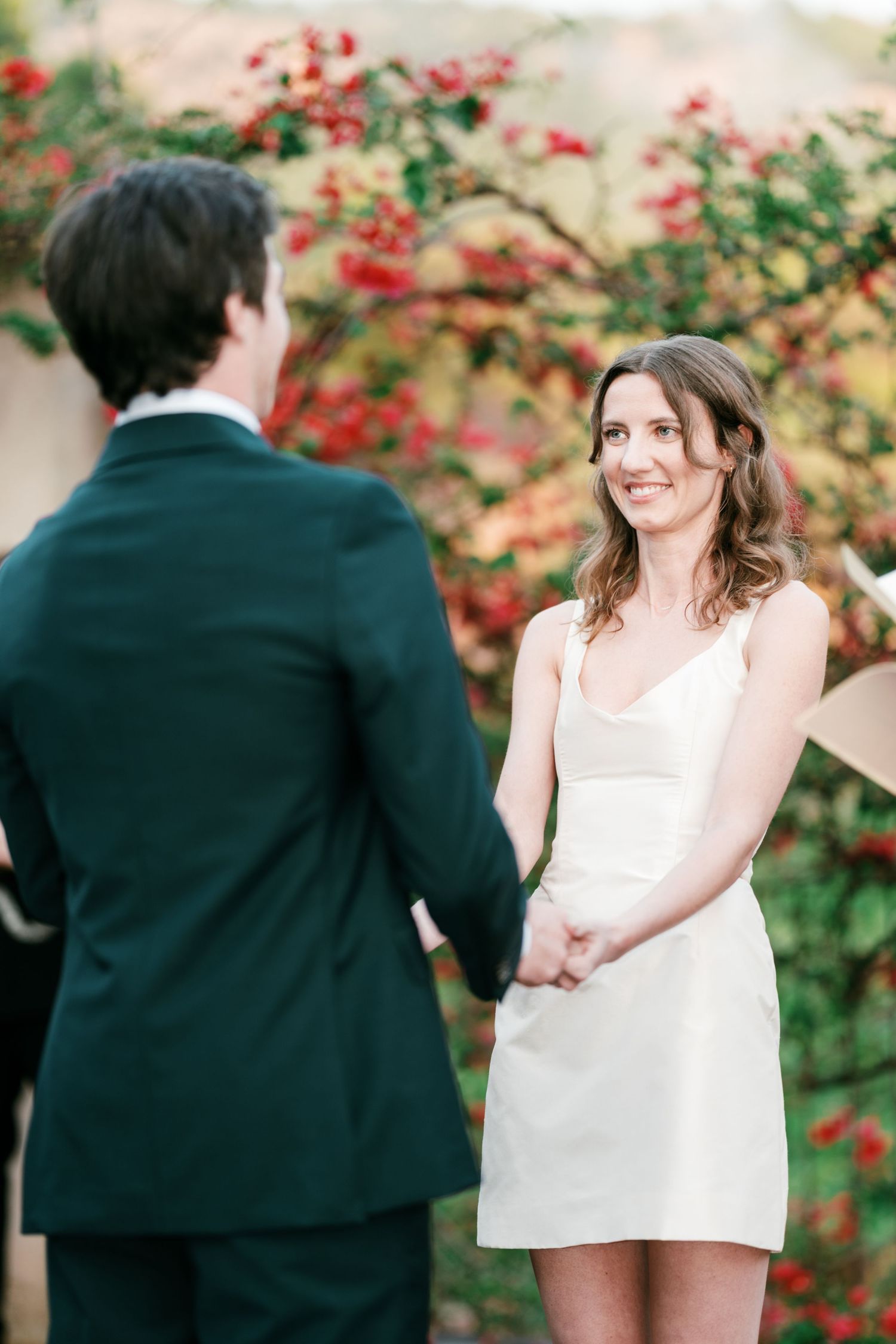 Groom in a dark suit holds hands with a smiling bride in a white dress in front of blooming red flowers.