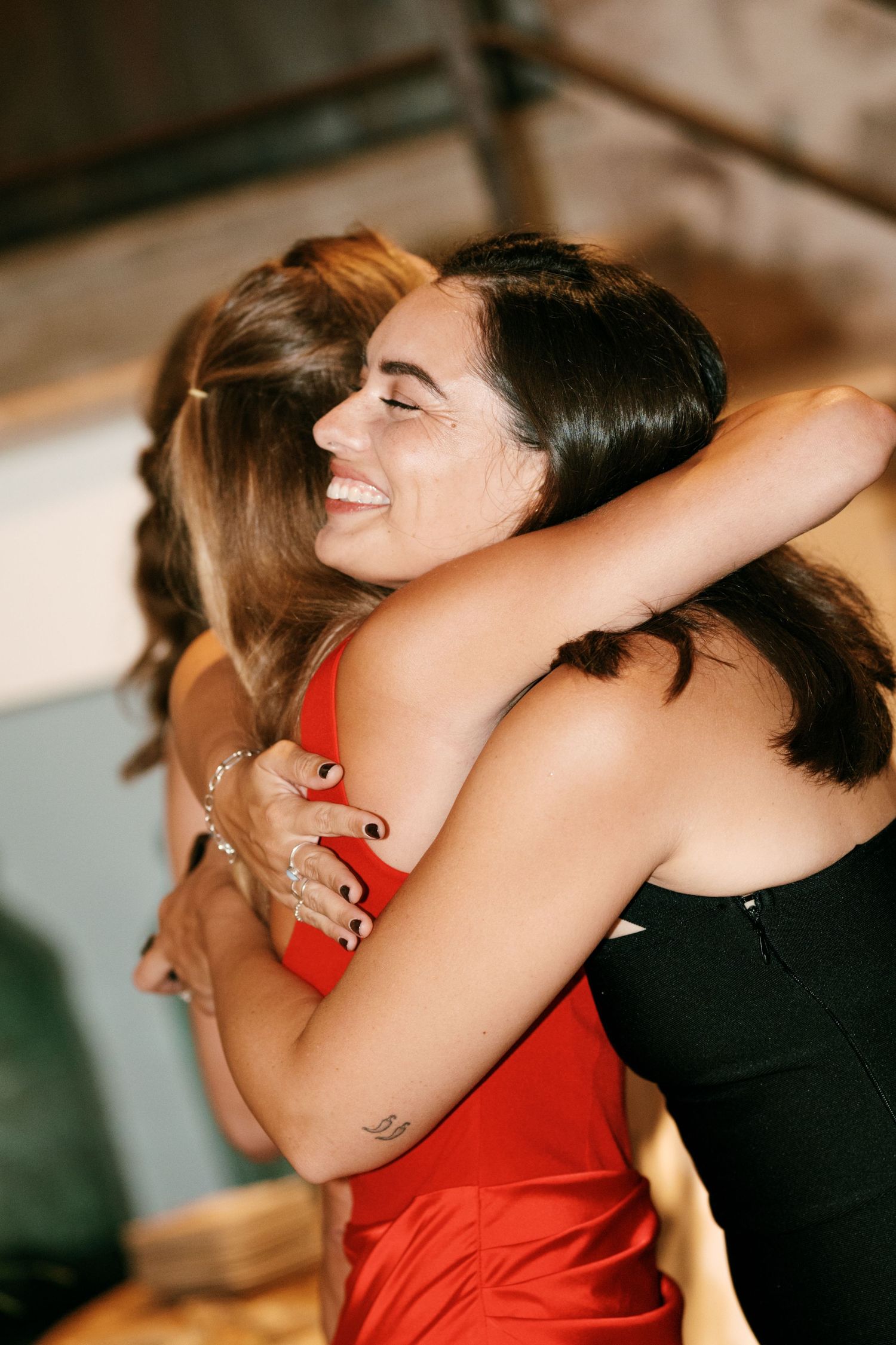 Two smiling women in red and black dresses share a warm embrace at a festive indoor celebration.