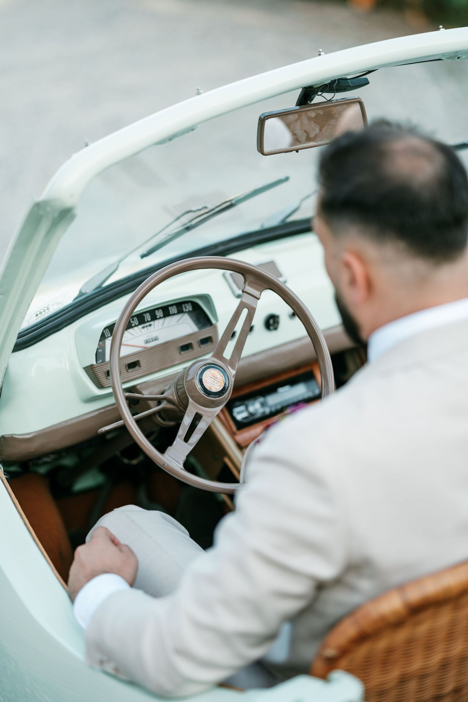 Man in a beige linen suit sits at the wooden steering wheel of a vintage cream convertible car with a wicker seat.