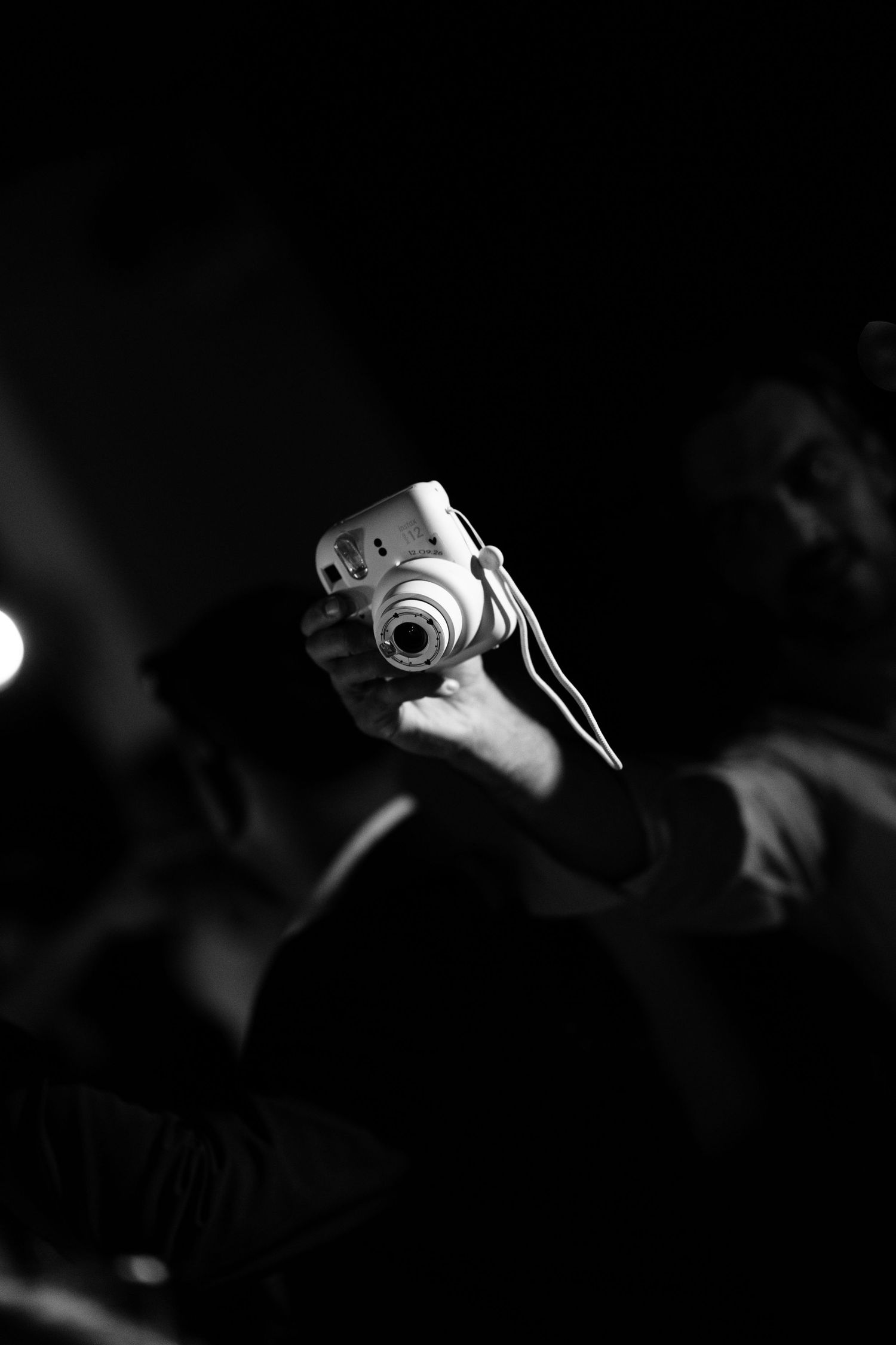 Black and white photo of a hand holding up a compact instant camera with a wrist strap, as blurred people stand in a dark room.