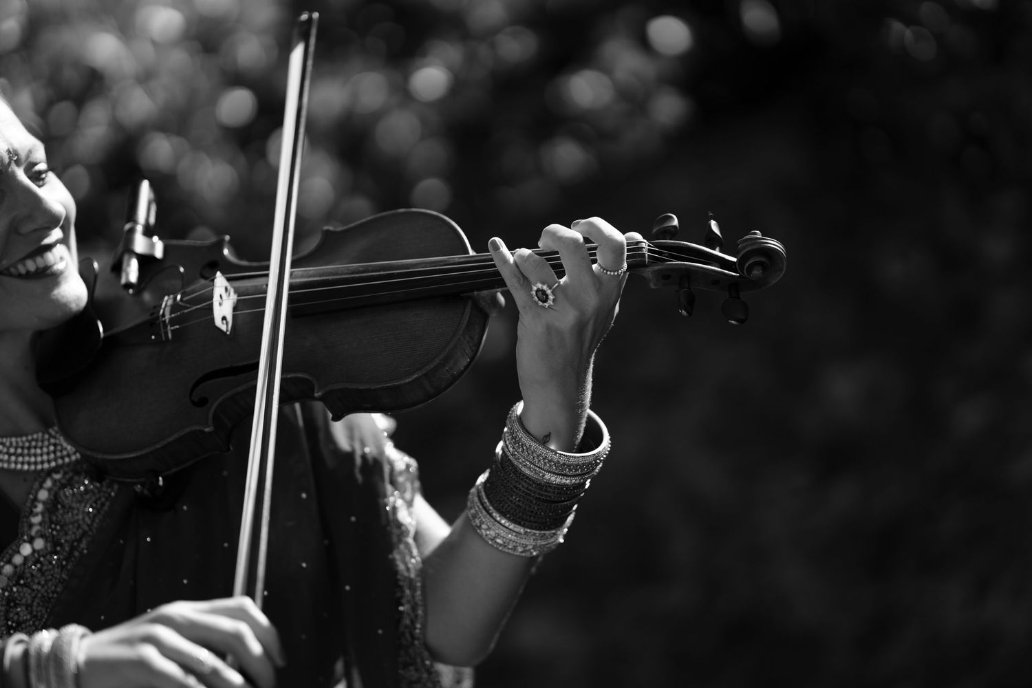Black and white photo of a smiling violinist wearing bangles and a ring, playing the violin outdoors with a soft blurred background.