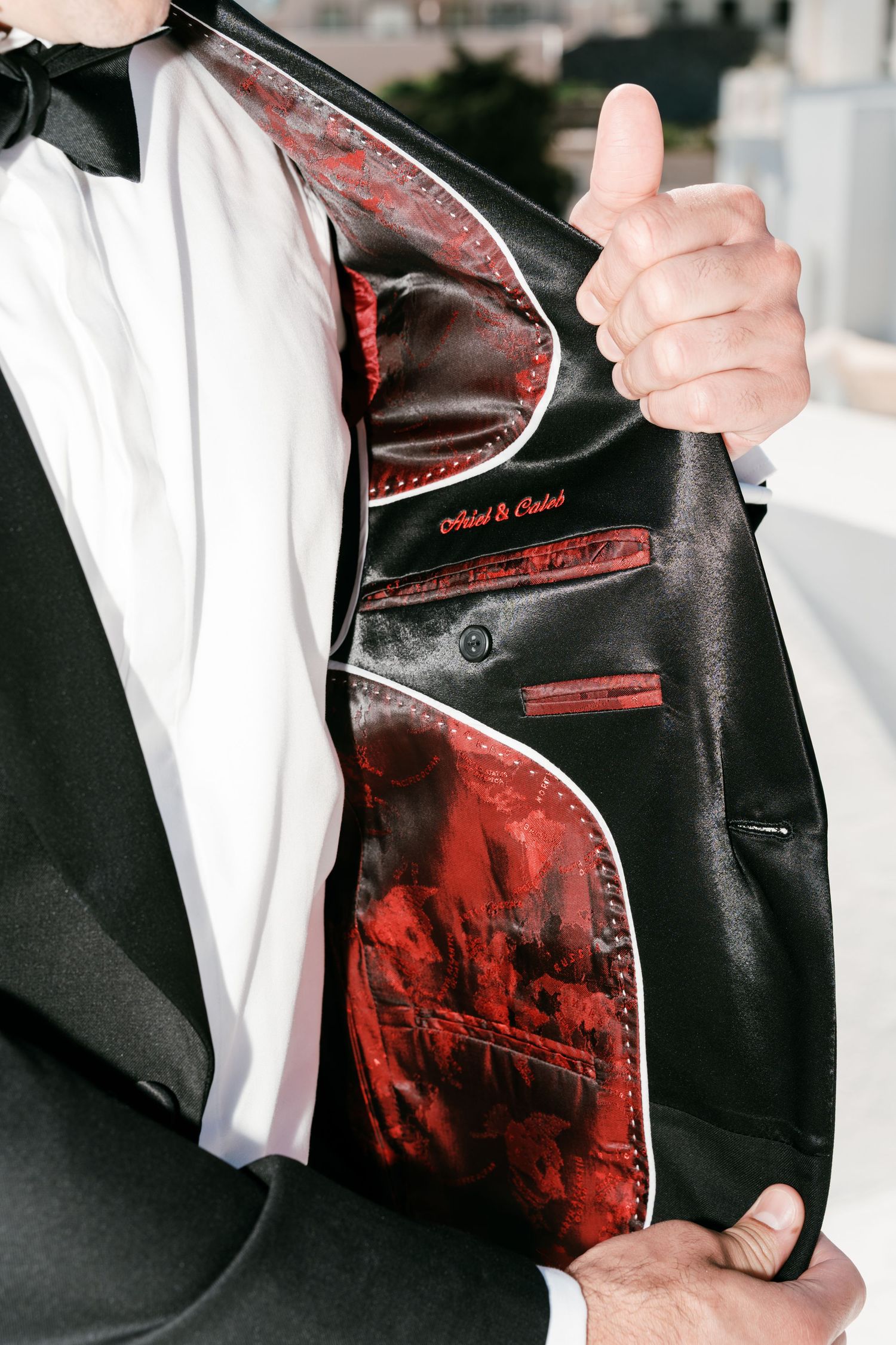 Man in black tuxedo displaying ornate red and black satin lining of jacket with thumbs up gesture.