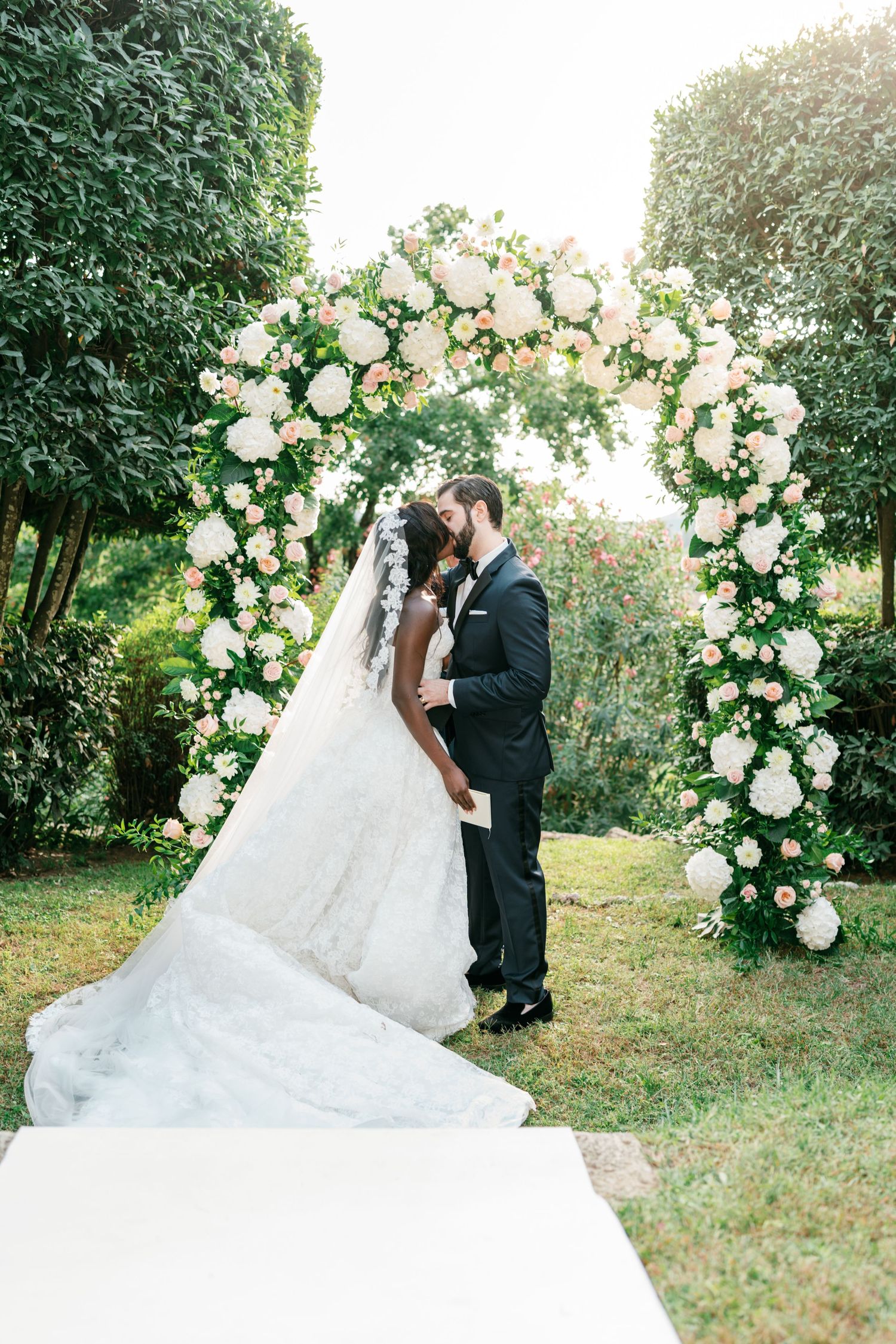Bride in a lace gown with a long veil kisses her groom in a black suit beneath a romantic floral wedding arch of white and blush roses.