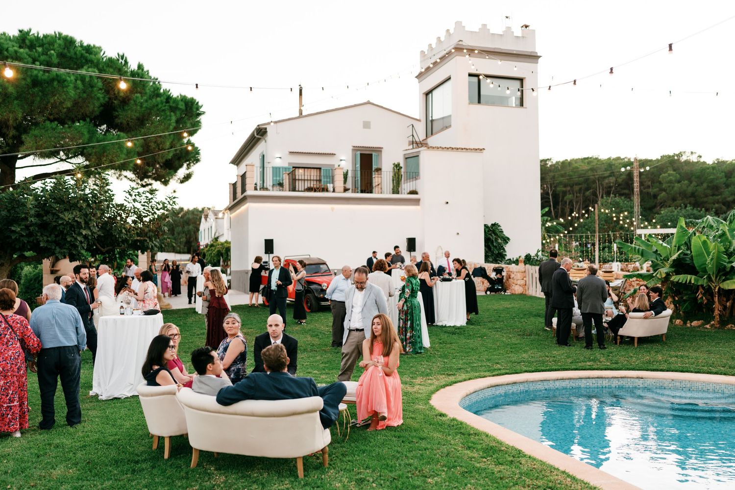 Guests mingle on a green lawn beside a swimming pool at an outdoor wedding reception in front of a whitewashed villa with a tower.