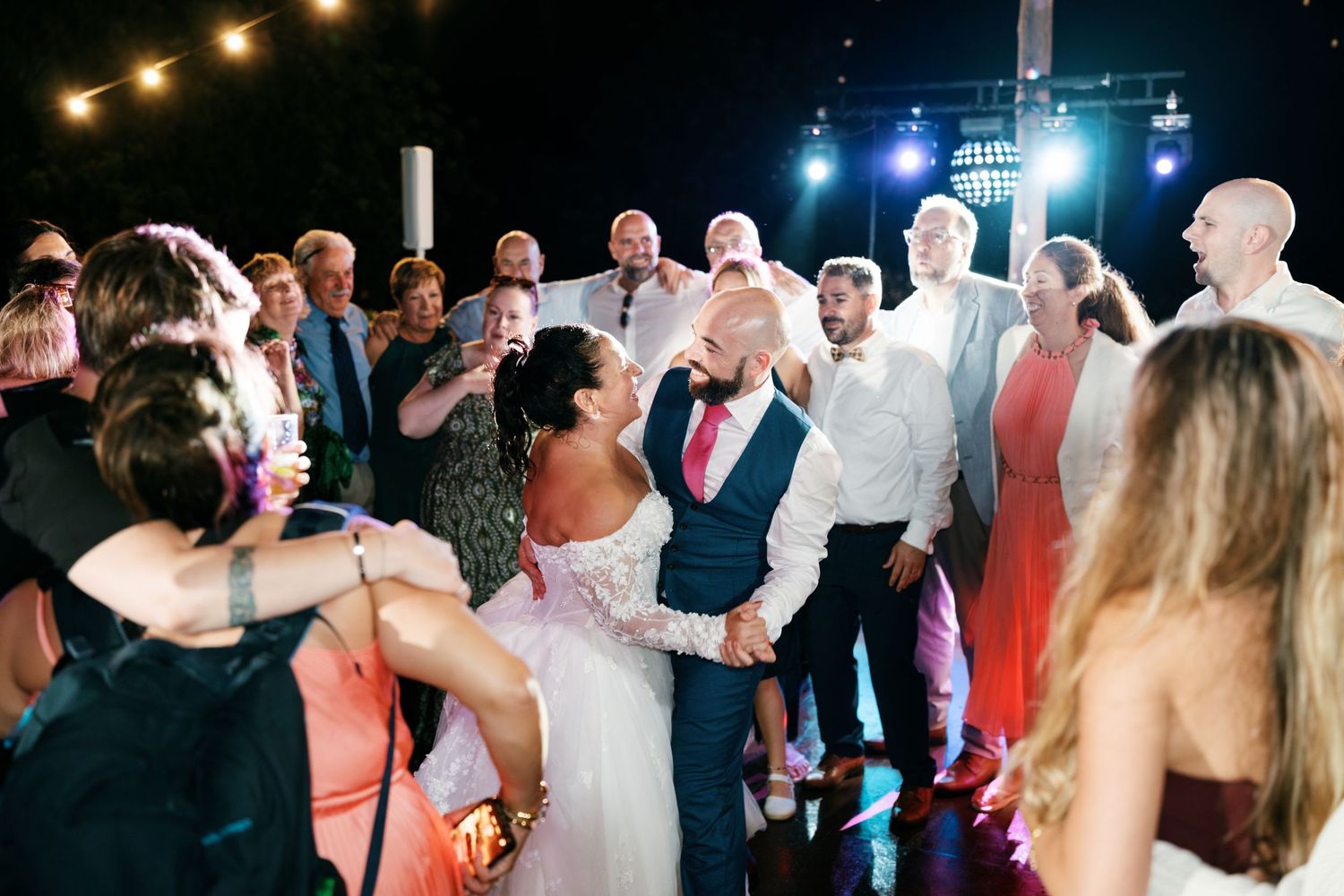 Bride and groom dance joyfully on the dance floor as smiling wedding guests cheer and clap around them at an evening reception.