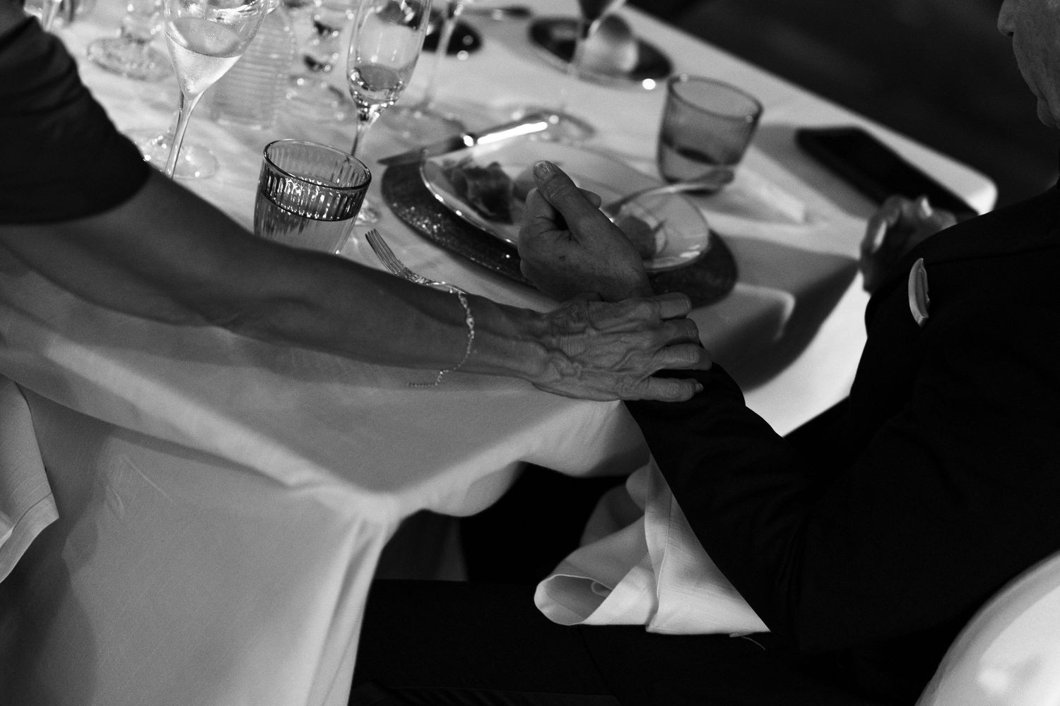 Black and white photo of two people at a dinner table with wine glasses, a plate of food, and hands resting on a white tablecloth.