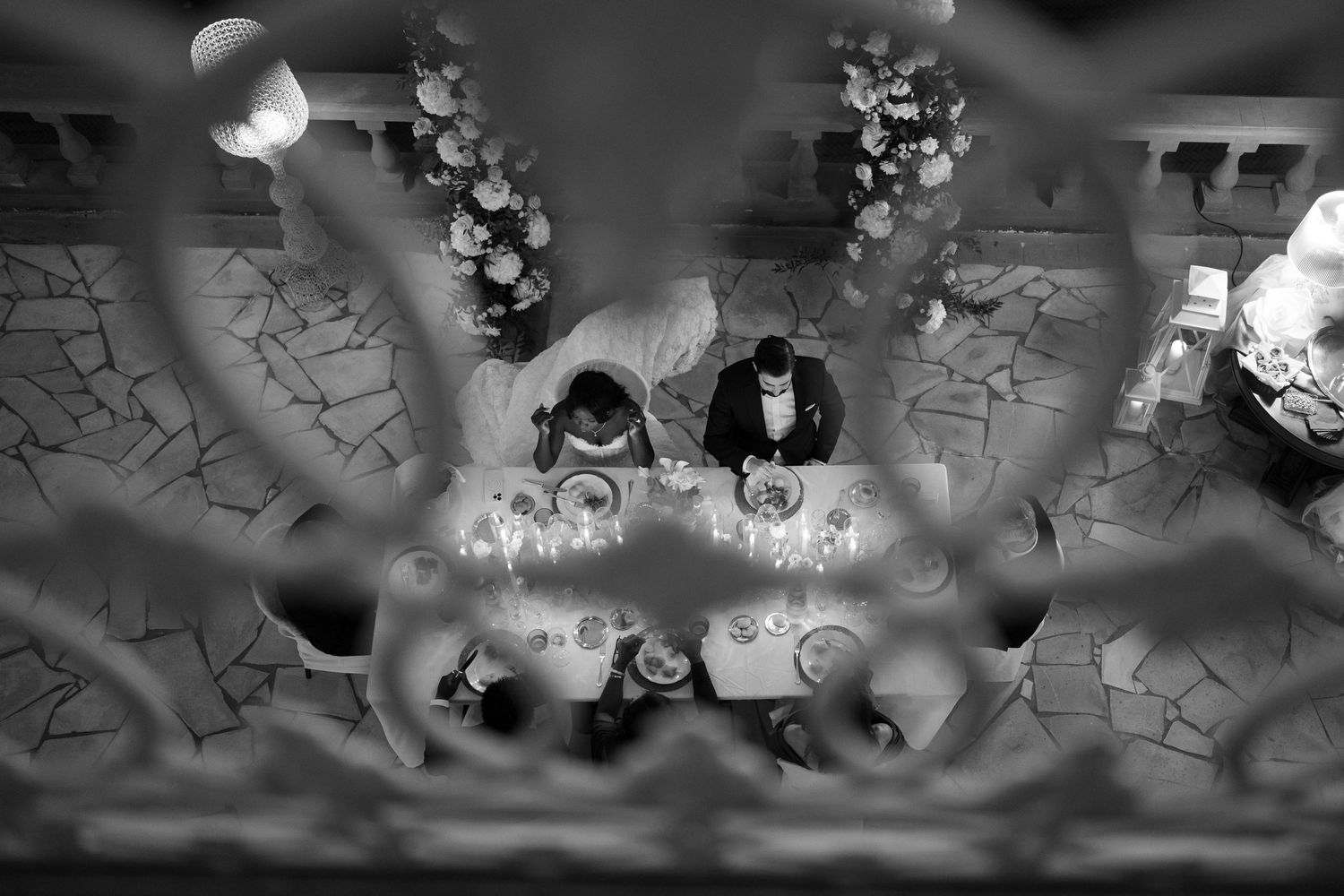 Black and white overhead view through ornate ironwork of a couple dining at a candlelit table with floral arrangements.