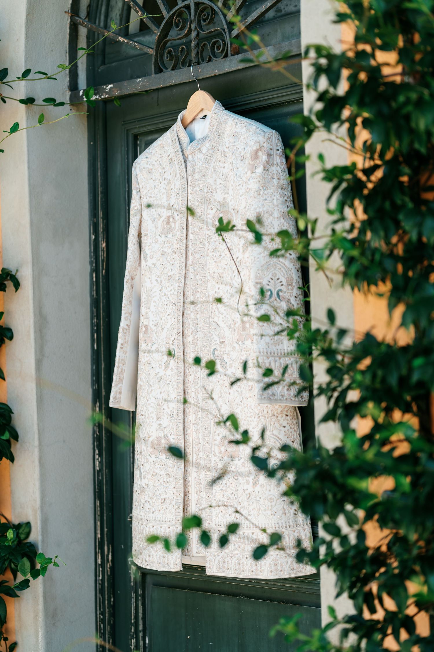 Ivory embroidered sherwani on a wooden hanger hanging from a dark green door, framed by leafy green vines.