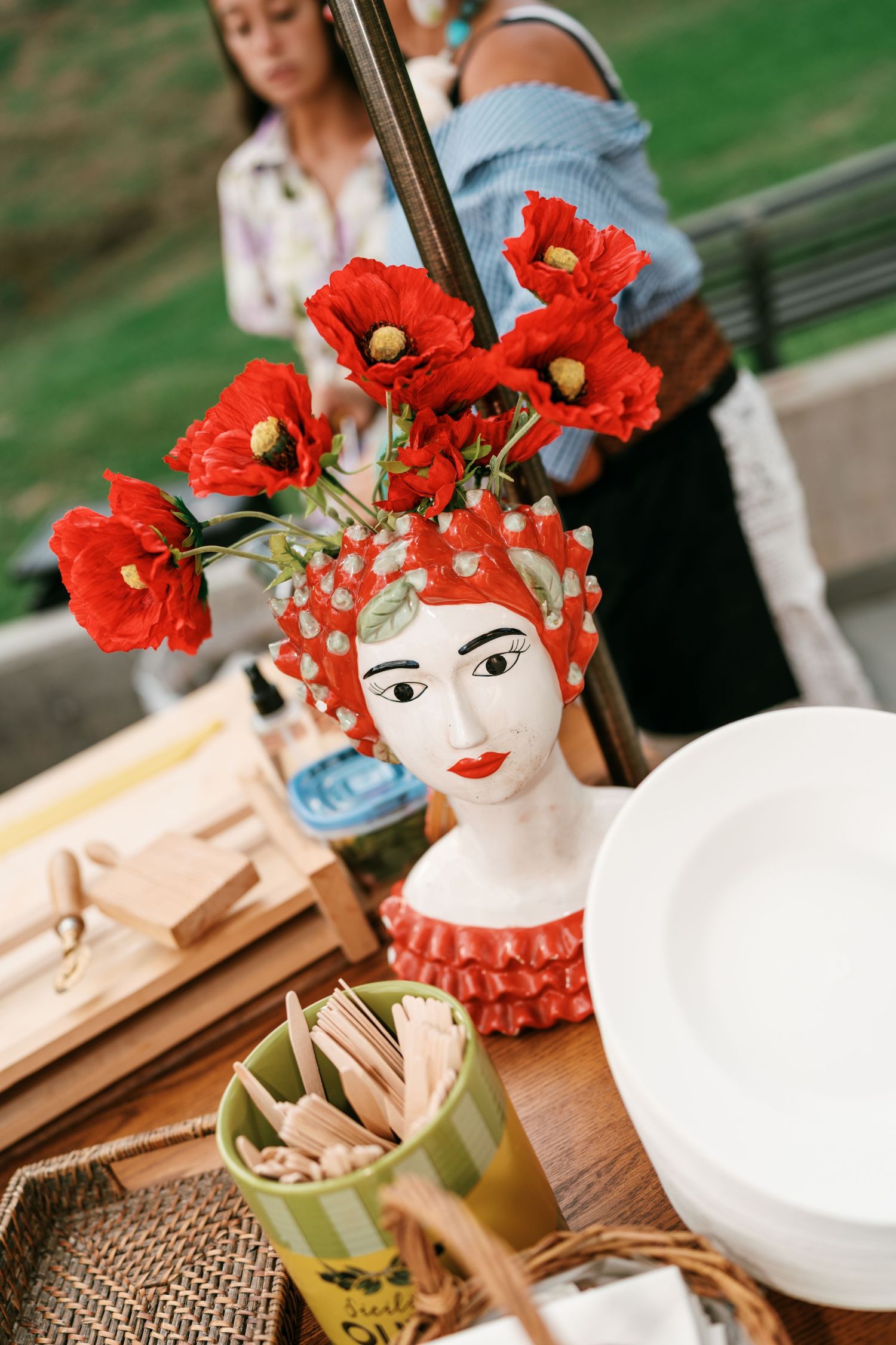 Ceramic woman's head vase with red poppies on a rustic outdoor table beside stacked white plates and wooden utensils.