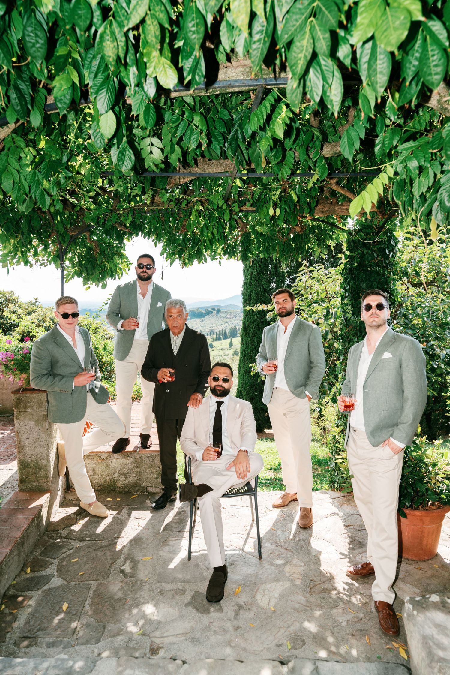 Six groomsmen in light suits and sunglasses hold drinks under a leafy vine-covered pergola at an Italian countryside wedding venue.