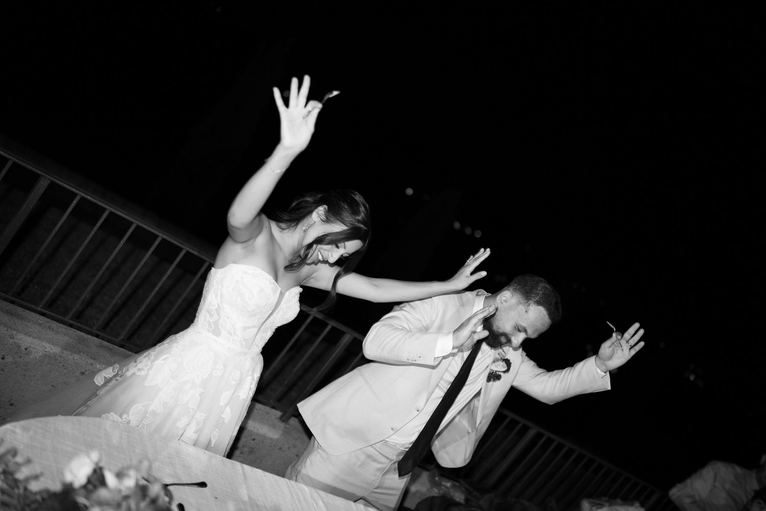Black and white photo of a bride in a white gown and a groom in a light suit dancing joyfully with raised arms at a night wedding reception.