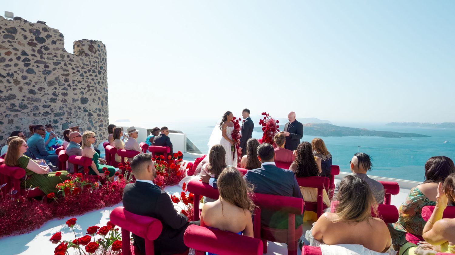Outdoor wedding ceremony in Santorini with red chairs, ocean views, and a couple exchanging vows near a stone tower.