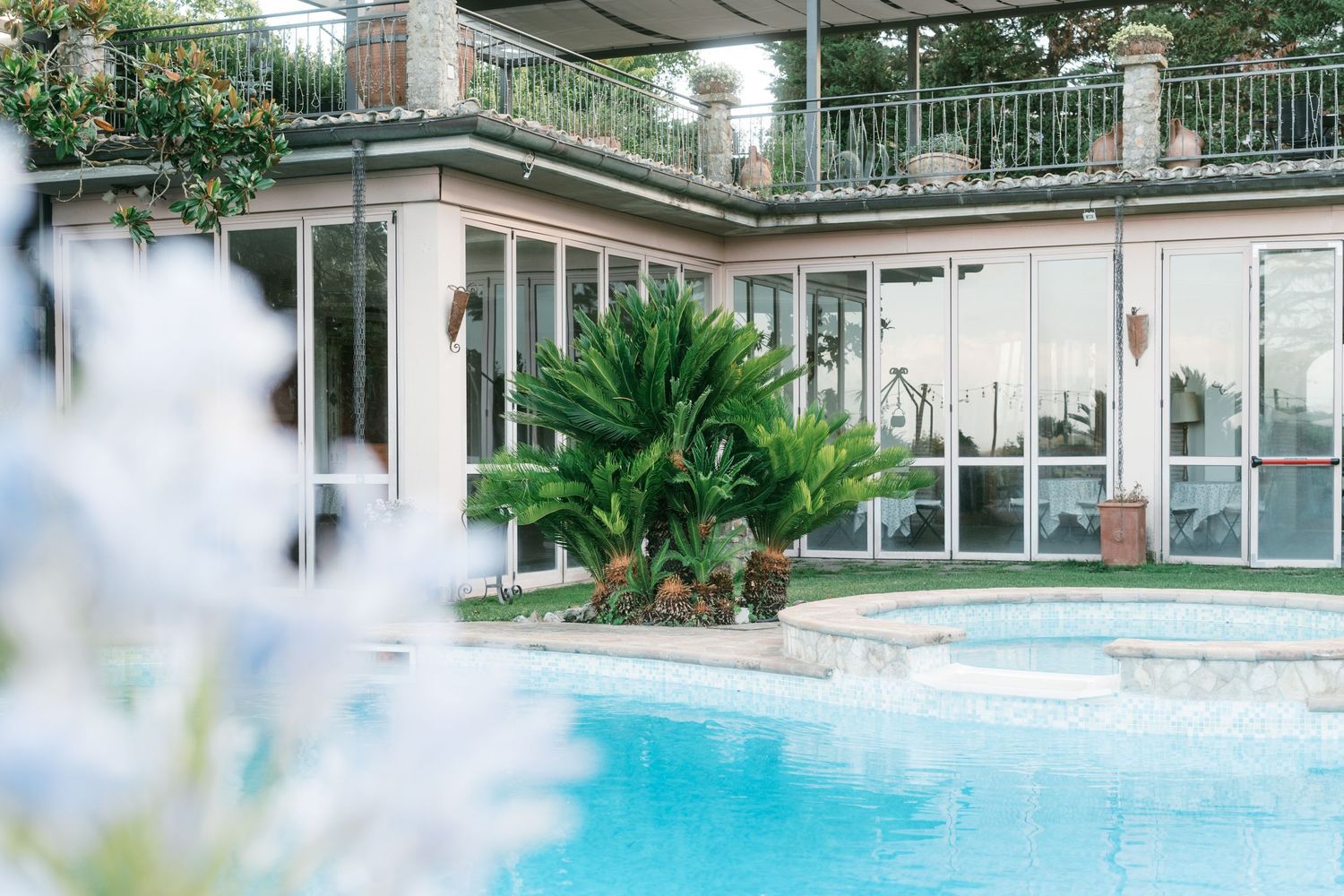 Elegant outdoor pool area with tropical palm trees and glass-windowed villa in background. Tuscany wedding photographer, filmmaker