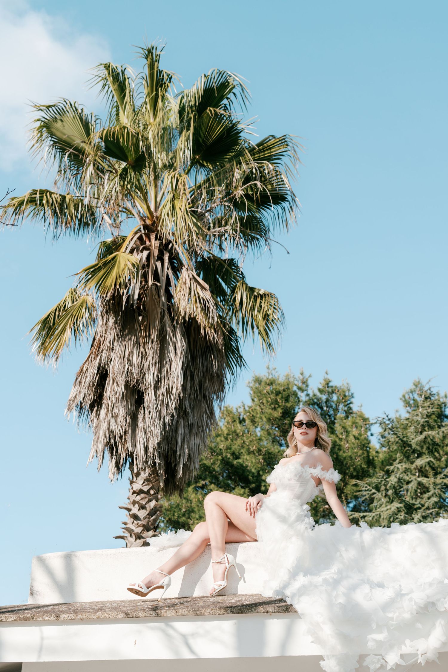 Blonde bride in white ruffled gown sits on white wall with palm tree and blue sky behind. Puglia wedding photographer, film