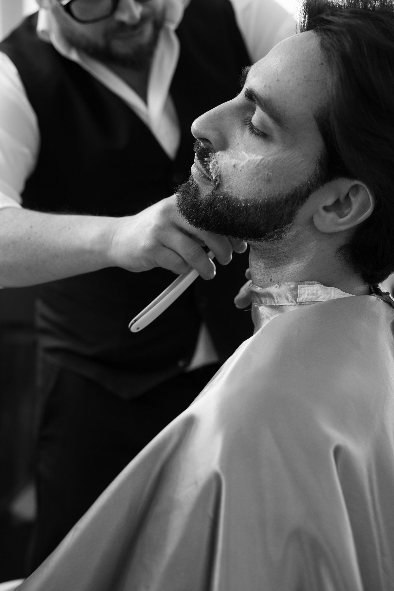 Black and white photo of a barber shaving a grooms beard with a straight razor. Puglia wedding photographer, film