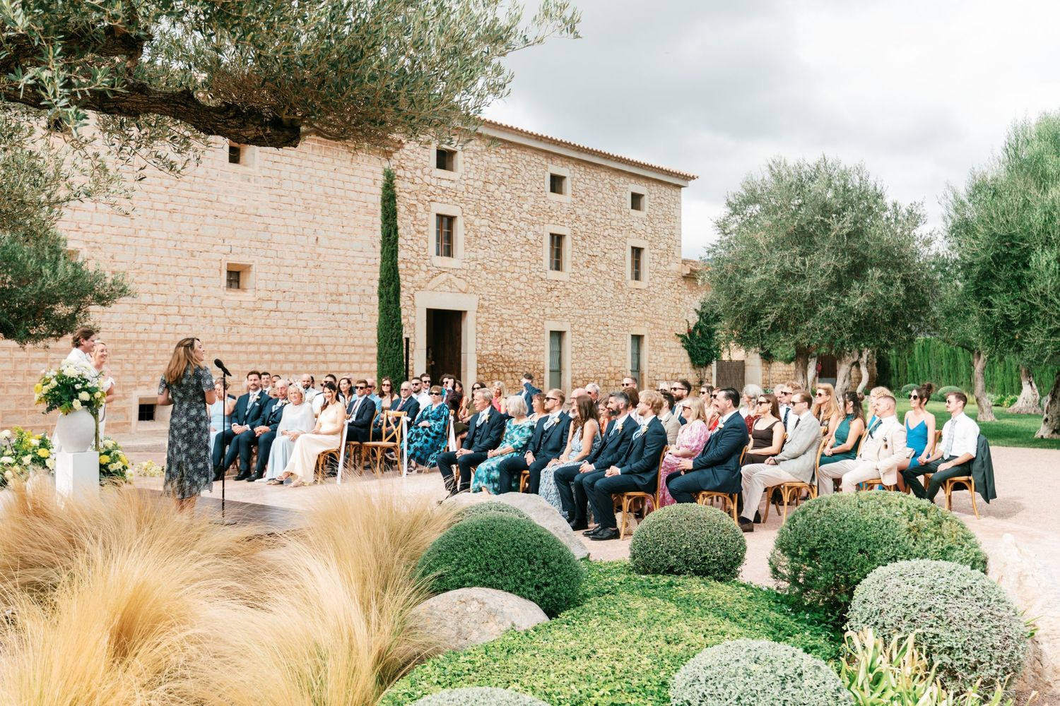 Wedding guests seated outdoors watch an outdoor ceremony in front of a rustic stone building, surrounded by olive trees, boxwood shrubs and 